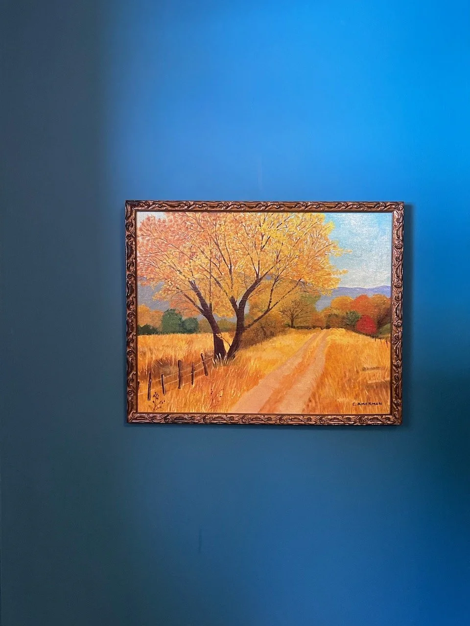 Painting of an autumn landscape with trees, a dirt path, and fields, framed and hanging on a blue wall.
