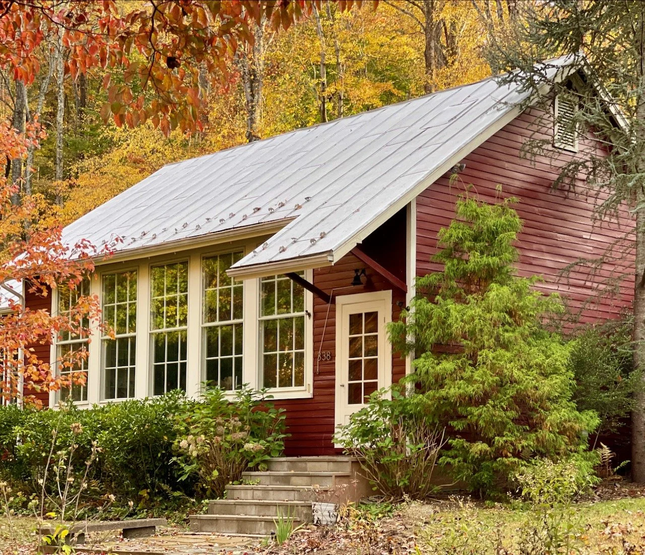 A red wooden house with white window frames and a gray metal roof, surrounded by green bushes and trees with fall foliage.
