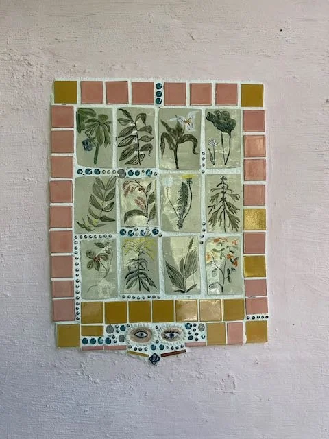 Decorative mosaic wall art featuring various botanical illustrations, surrounded by pink, yellow, and blue tiles, with a face at the bottom.
