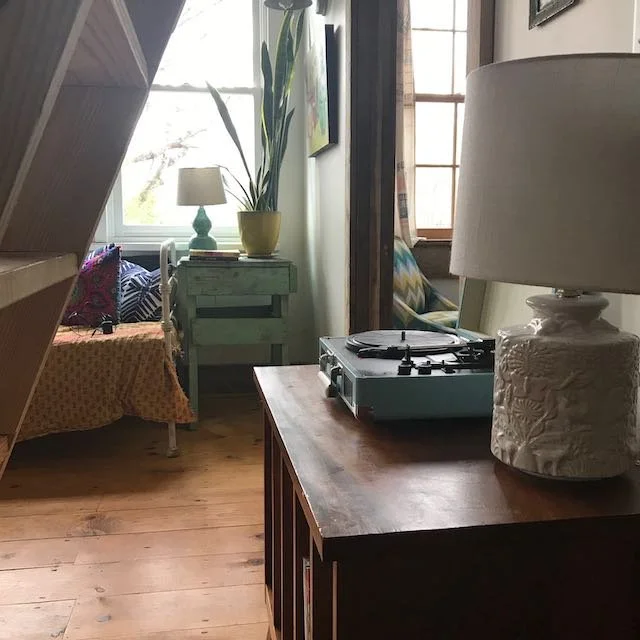 Living room with a record player on a wooden table, a lamp, a large potted plant, a window with curtains, and a seating area with cushions and a blanket.