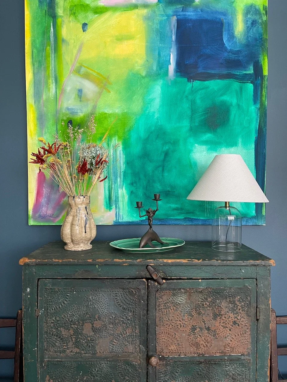 A decorative still life against a colorful abstract painting. The setup includes a vintage green cabinet with peeling paint, a ceramic vase with dried flowers, a black candlestick holder with three arms, and a glass table lamp with a white shade.