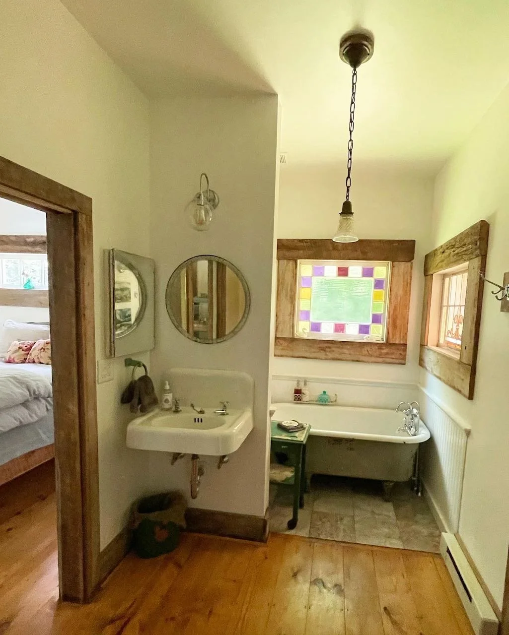 Interior view of a vintage-style bathroom with a small wall-mounted sink, round mirrors, a clawfoot bathtub, and stained glass window, featuring wooden accents and rustic decor.