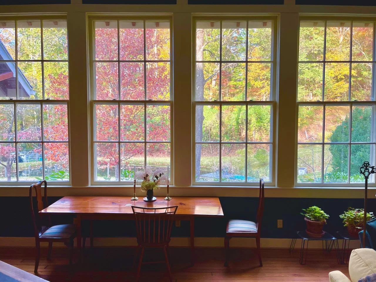 Interior view of a dining room with three large windows showing colorful autumn trees outside. A wooden table with three chairs, a flower arrangement, and two candlesticks are on the table. Potted plants are on a small stand near the right window.