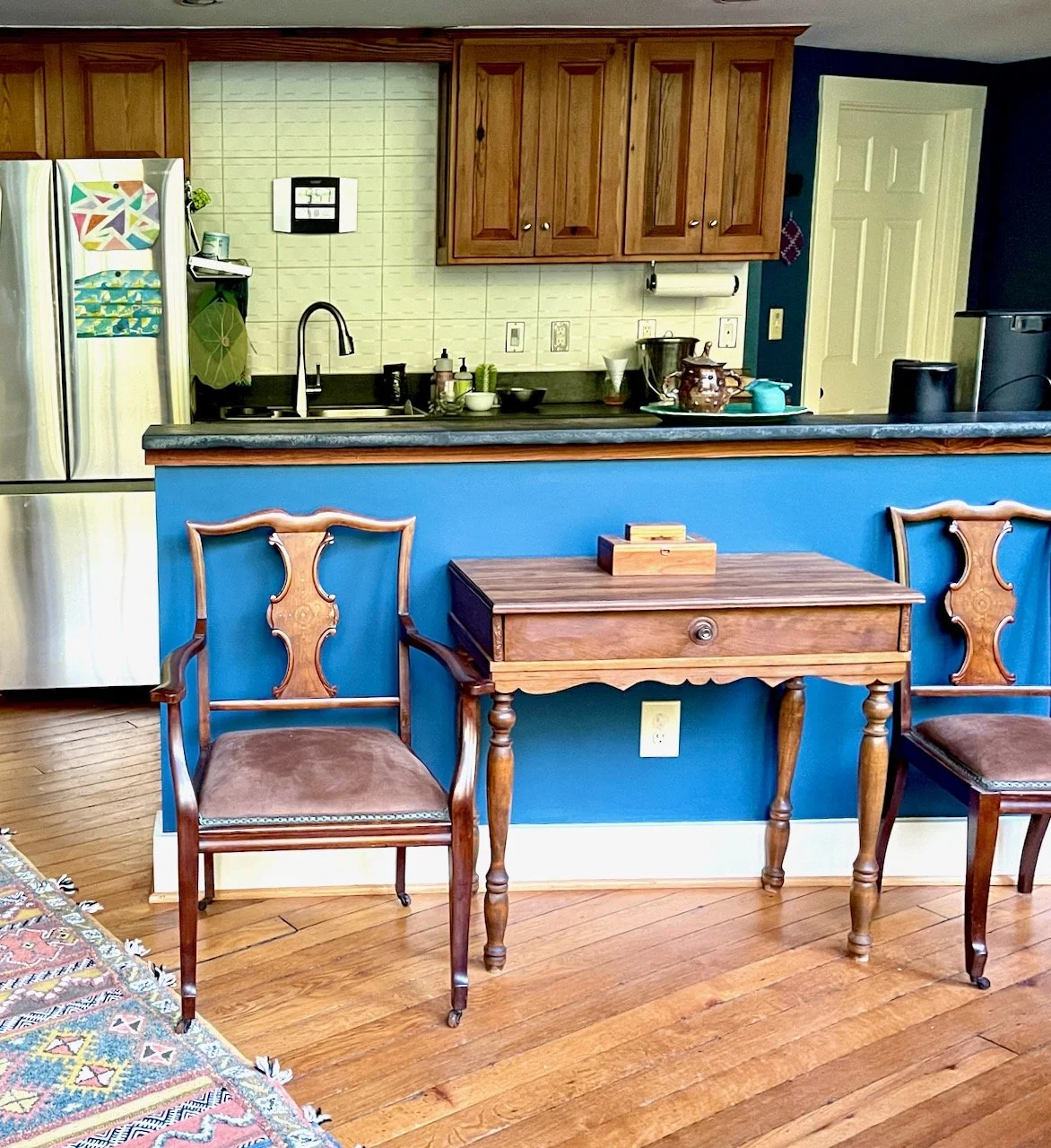 A kitchen with wooden cabinets, a black countertop, and a white tiled wall. There is a stainless steel refrigerator with colorful magnets and a front laundry message. A blue breakfast bar separates the kitchen from the dining area, which has two wood