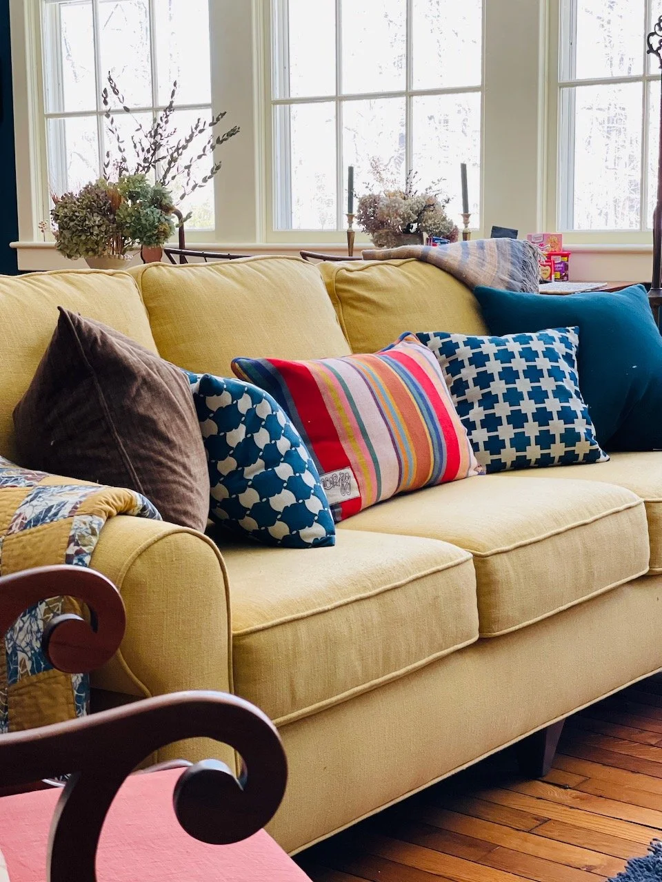 A yellow upholstered sofa with four decorative throw pillows in various patterns and colors, set in a room with large windows and wooden floors. Behind the sofa are plants and candles.