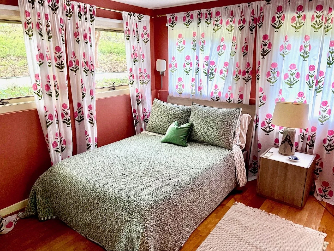 A cozy bedroom with a double bed covered in floral patterned bedding, surrounded by curtains with matching floral prints, a small wooden bedside table with a lamp, and a window showing greenery outside.