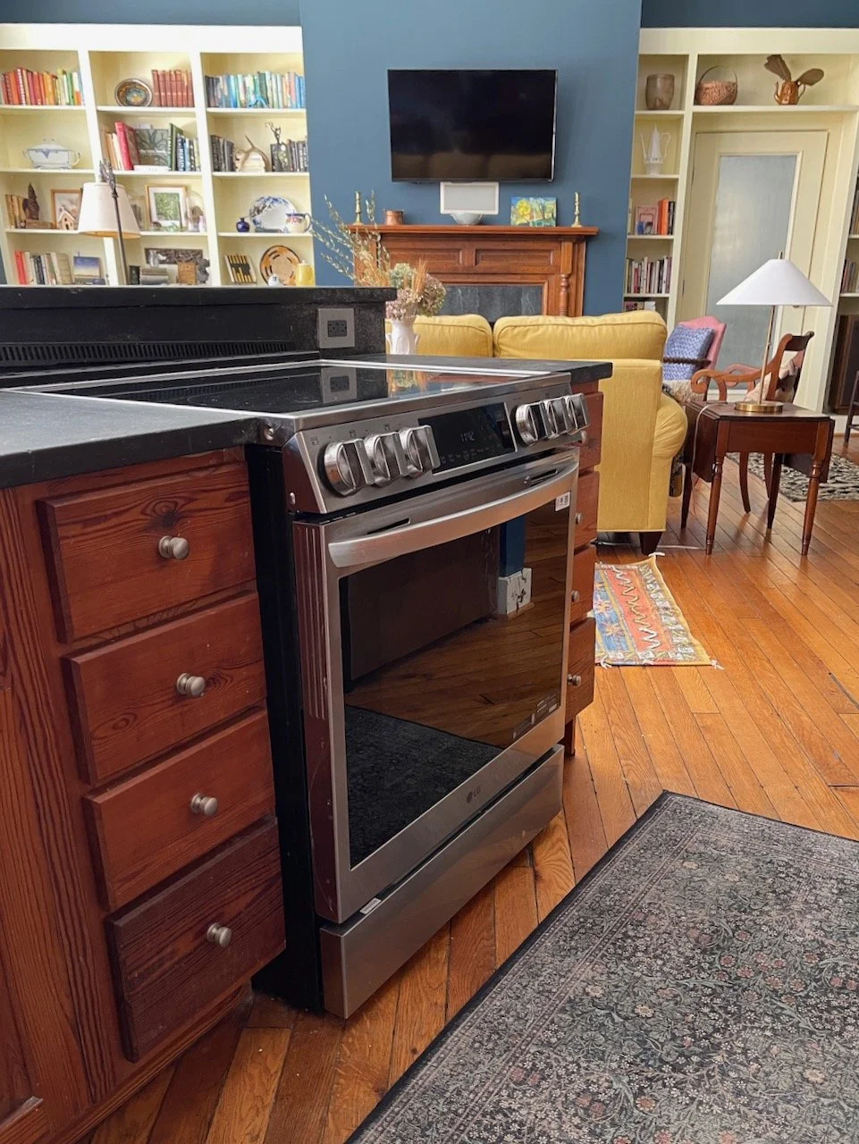 View of a kitchen with a stainless steel oven/stove, wooden cabinets, and a black countertop. In the background, there is a living room area with a yellow sofa, a navy blue accent wall with a mounted TV, a wooden fireplace mantle, bookshelves filled 