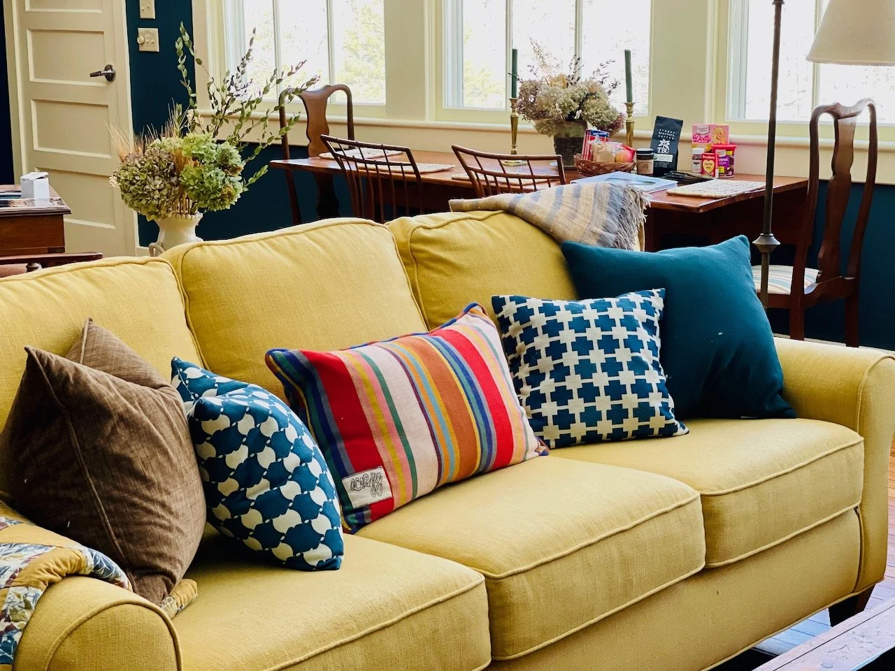 A living room with a yellow sofa decorated with colorful pillows, behind a wooden dining table with flower arrangements, windows, and a blue wall.