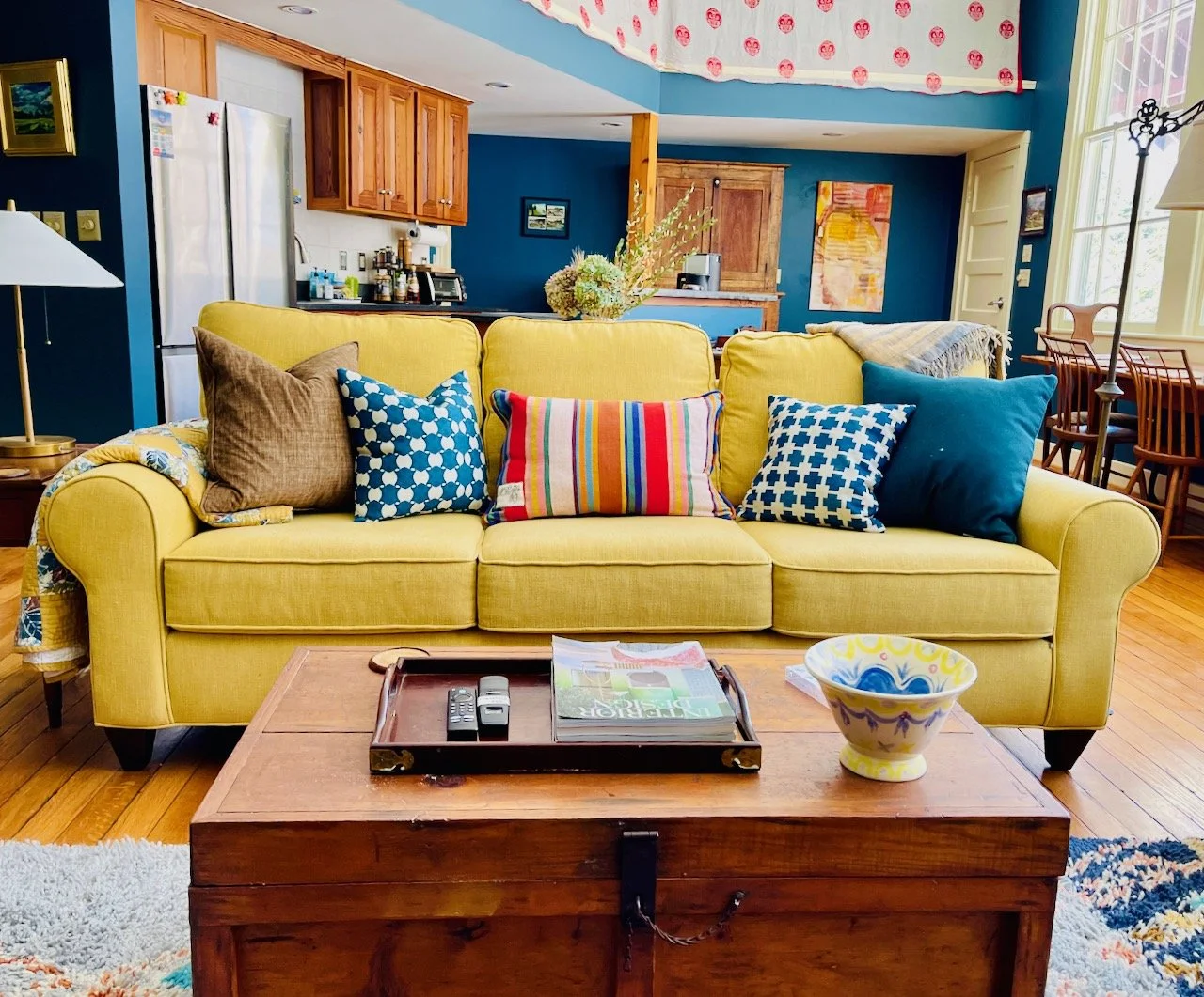 Living room with a yellow sofa, colorful pillows, wooden coffee table with magazines and a decorative bowl, blue walls, kitchen area in the background, and large windows.