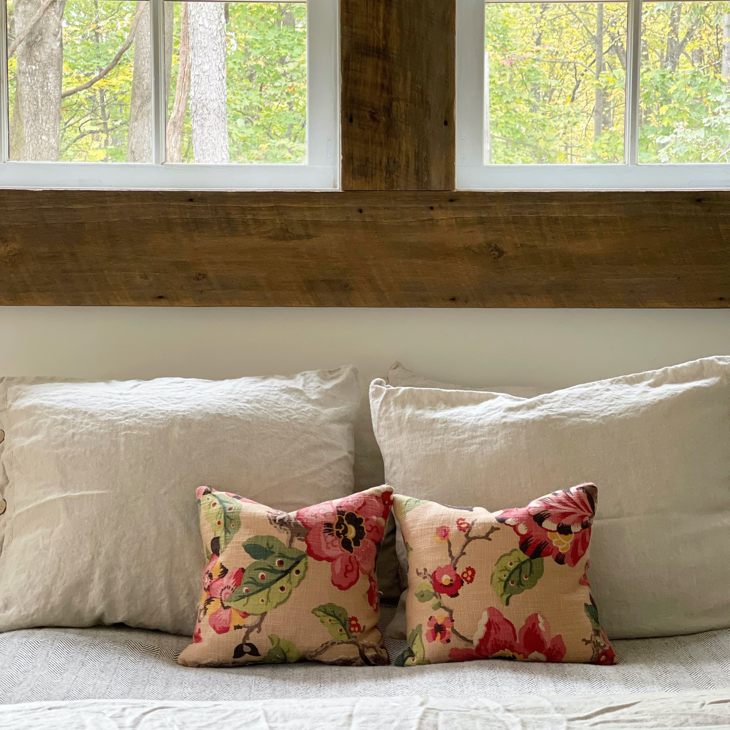 Close-up of a bed with three white pillows and two decorative floral pillows in front. Behind the bed is a window with a view of green trees.