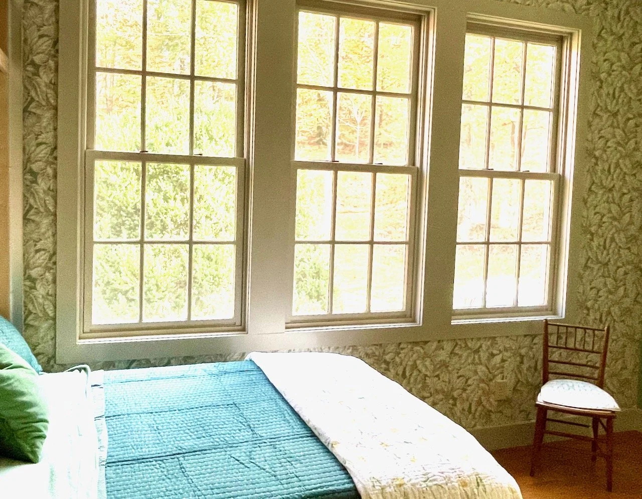Bedroom with three large windows letting in sunlight, a bed with blue bedding and a green pillow, patterned wallpaper, and a red wooden chair with a cushioned seat.