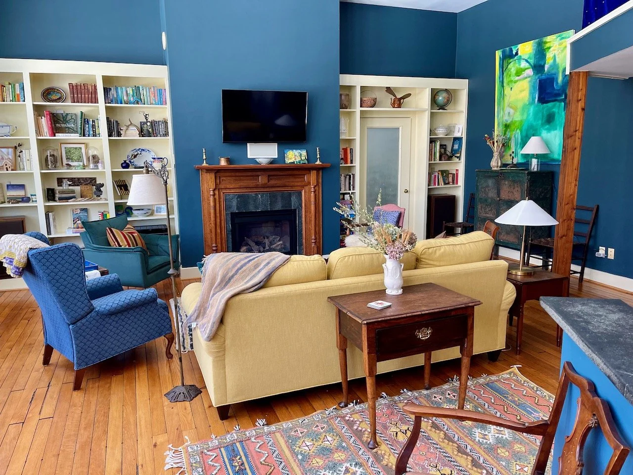 Living room with yellow sofa, blue armchairs, wooden flooring, bookshelf, fireplace with TV, and colorful wall art.