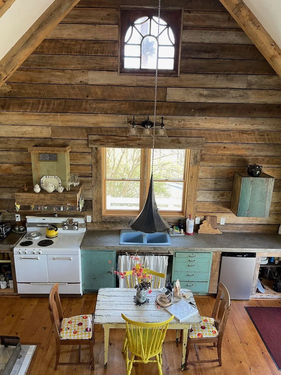 A rustic kitchen with wooden walls and ceiling, featuring a central window above a blue sink and a black hanging light fixture. There is a white stove on the left, teal cabinets, a small refrigerator, and a wooden dining table with four chairs, two o