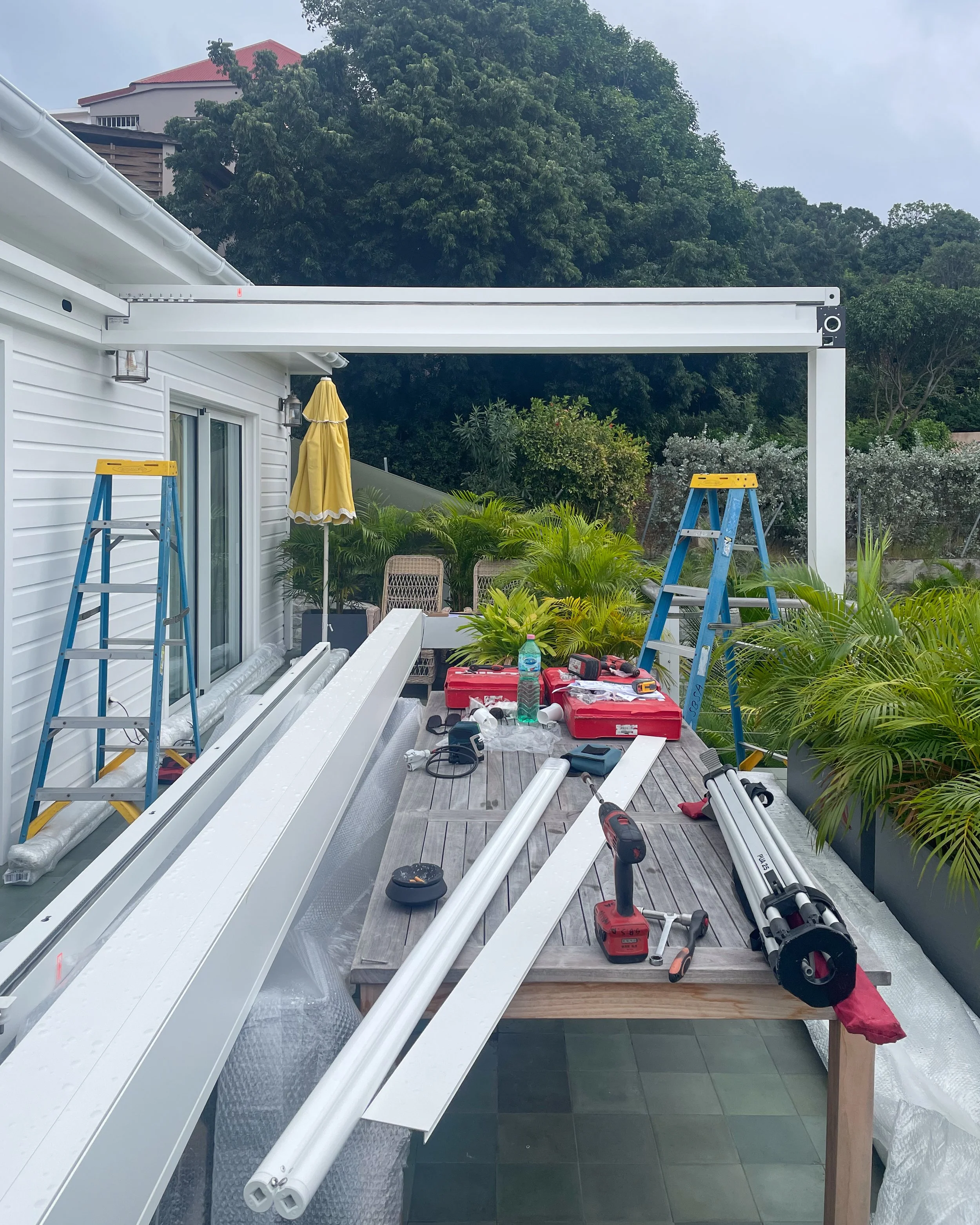 Outdoor workspace with tools and materials for building a pergola or canopy, including metal and plastic pipes, power tools, and ladders, set on a wooden table with greenery and a house in the background.