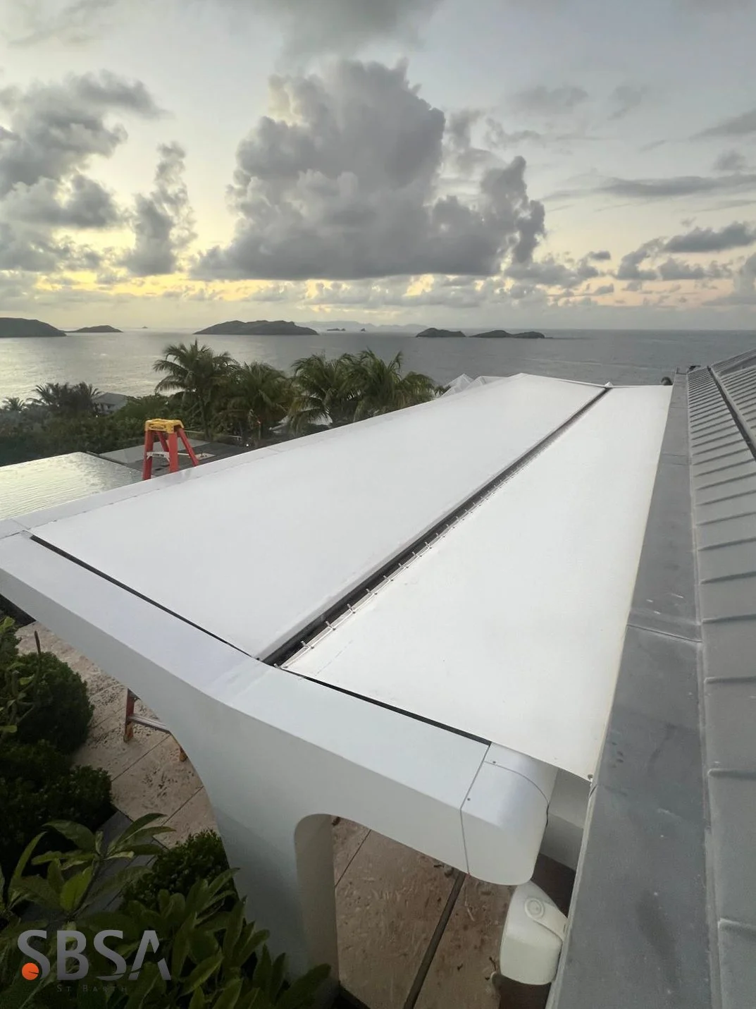 A white grand piano on a balcony overlooking the ocean with small islands in the distance, cloudy sky, and palm trees.