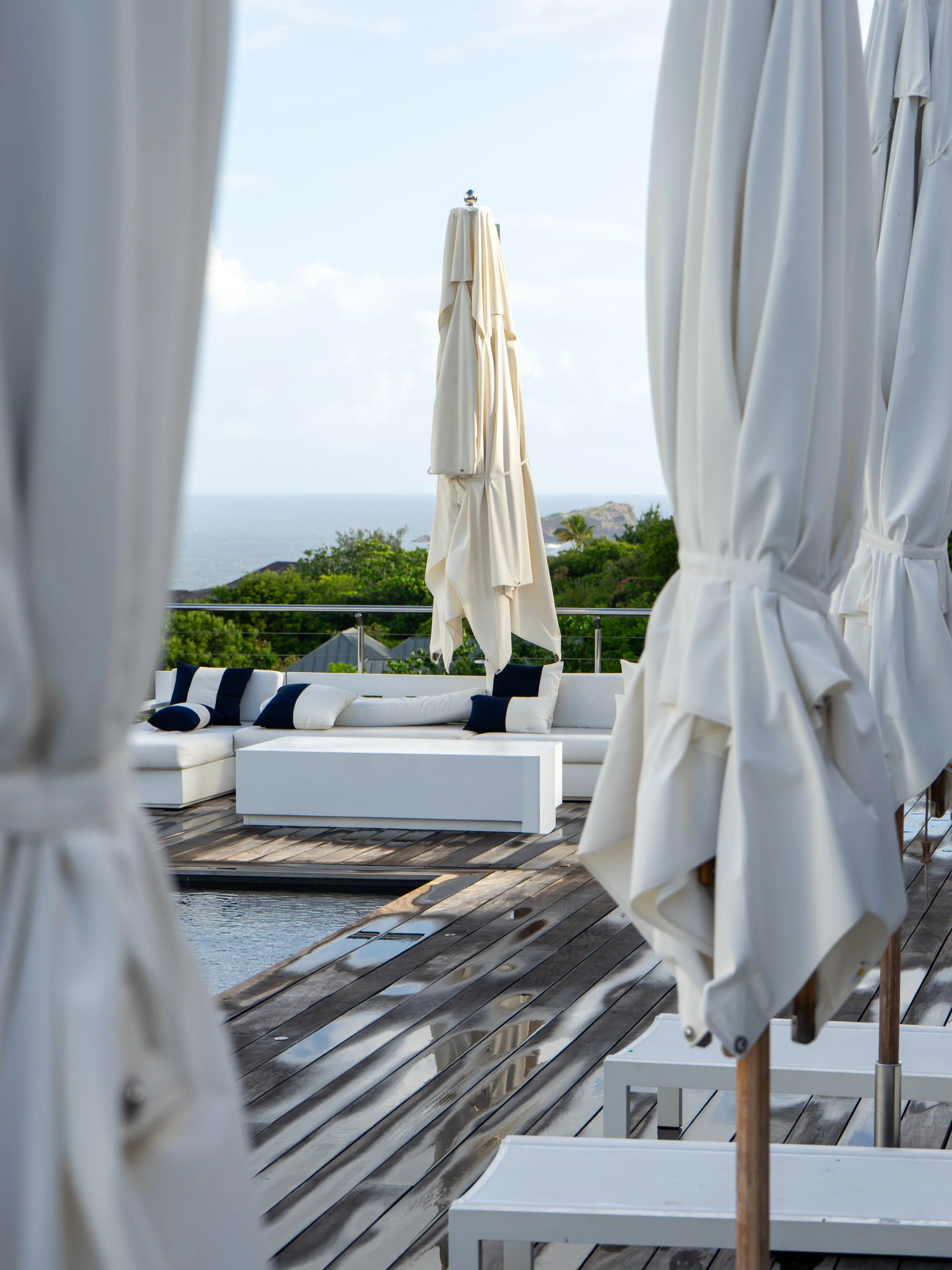 View of a seaside patio with white curtains on either side, a white outdoor sofa with black and white pillows, a wooden deck, and a large closed white umbrella, overlooking the ocean and green landscape.