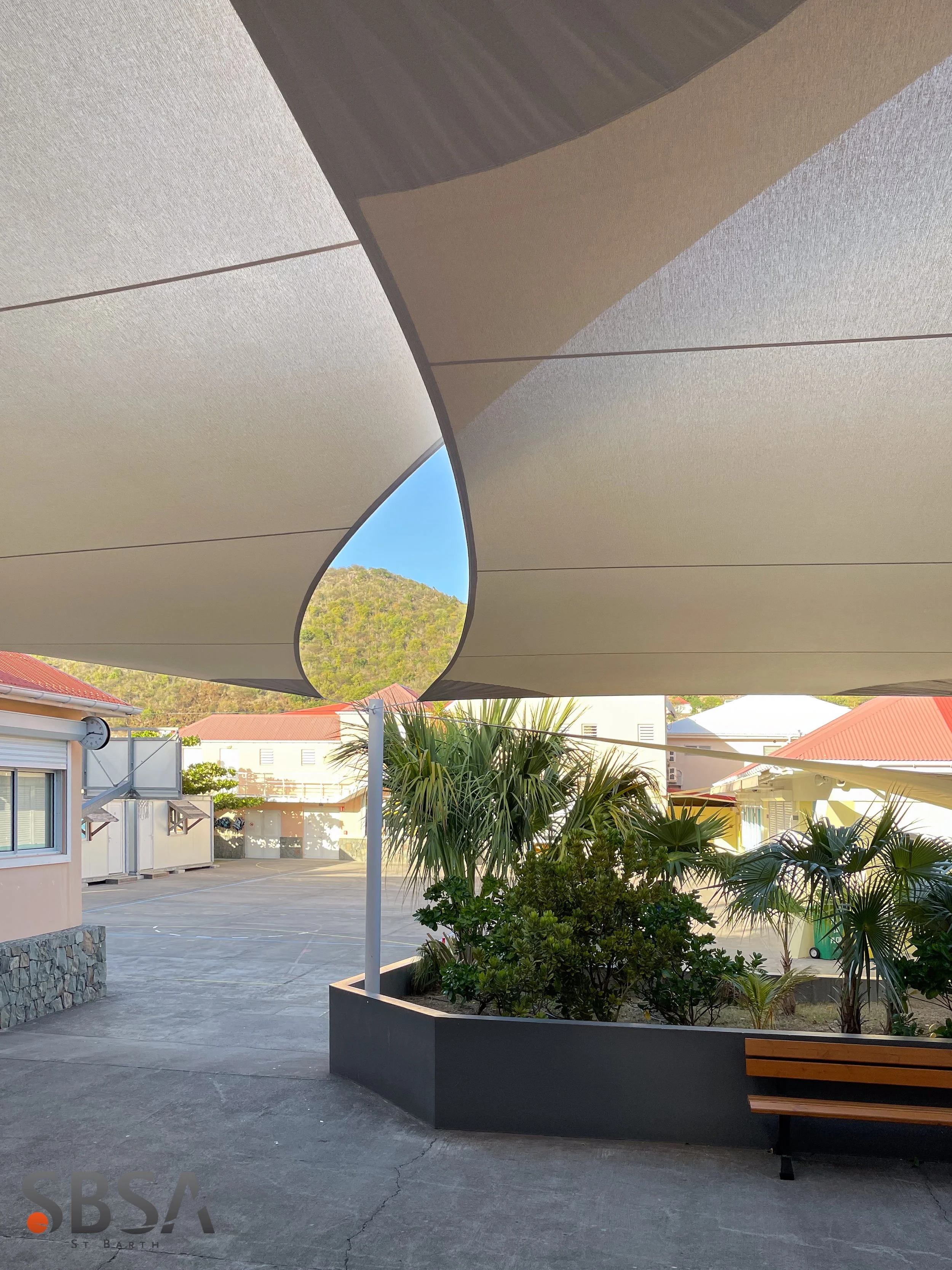 Outdoor area with modern shaded structure, benches, a few small trees, a building with a clock, parking lot, and distant hills