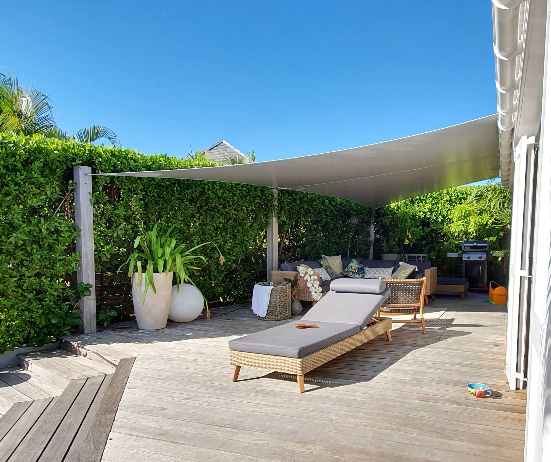 Decorated outdoor patio area with wooden decking, a sunshade, outdoor couches with cushions, large white planters with green plants, and a bright blue sky.