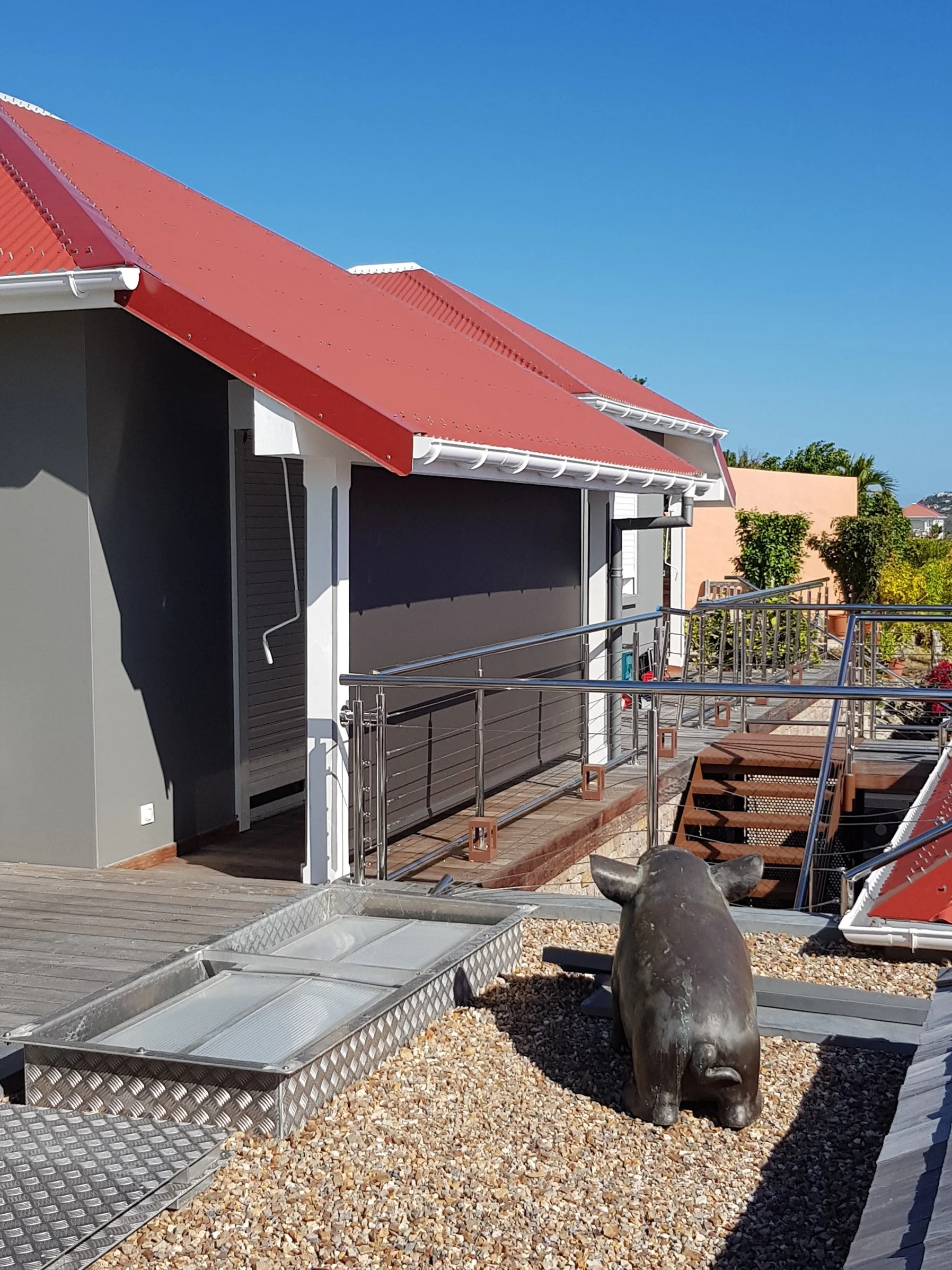 A house with a red metal roof, a dark gray wall, a balcony with stainless steel railing, and a decorative sculpture of a pig. The ground is covered with small rocks, and there are gardening supplies and tools nearby. The sky is clear and blue.