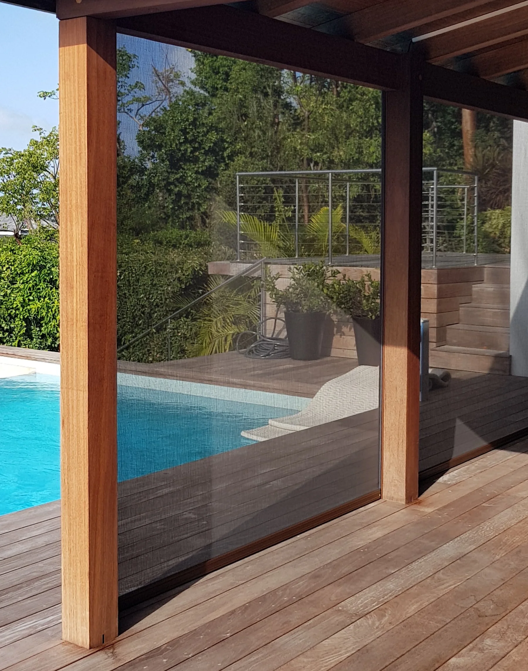 View of a backyard pool area through a glass door with a wooden frame and screen, showing a pool, potted plants, a wooden deck, a privacy fence, and green trees and bushes in the background.