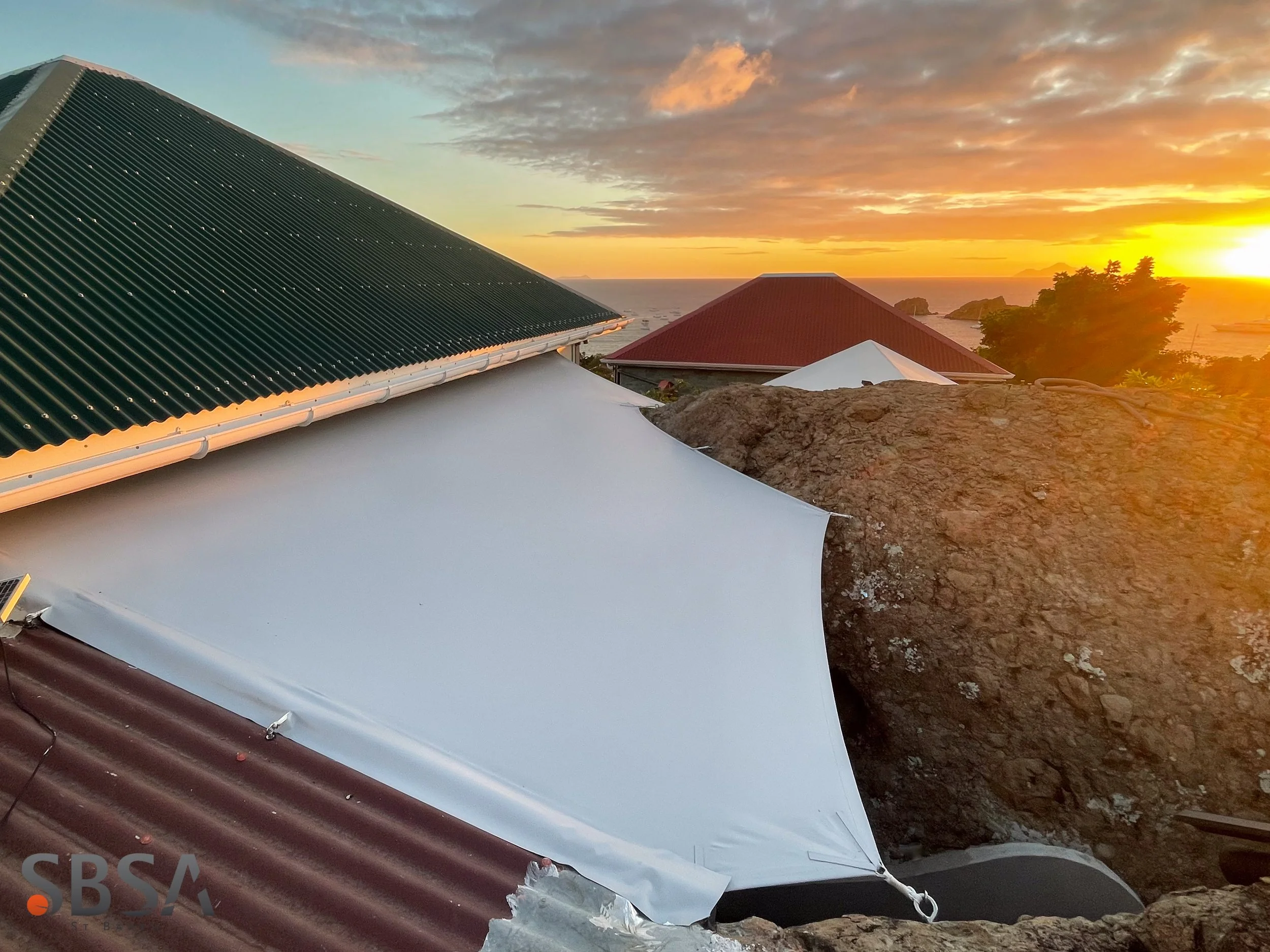 Sunset over rooftops with stormy clouds and ocean in the background, some houses with red, white, and green roofs