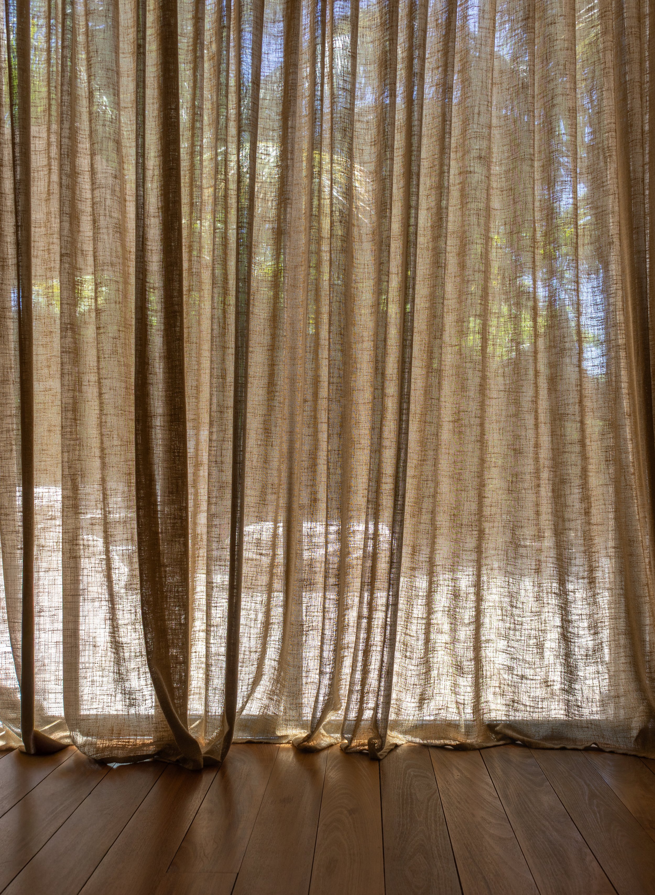 Des rideaux semi-transparents de couleur claire sont suspendus devant une fenêtre laissant filtrer la lumière du soleil, avec une vue sur des arbres à l’extérieur et un parquet en bois visible au bas de l’image.