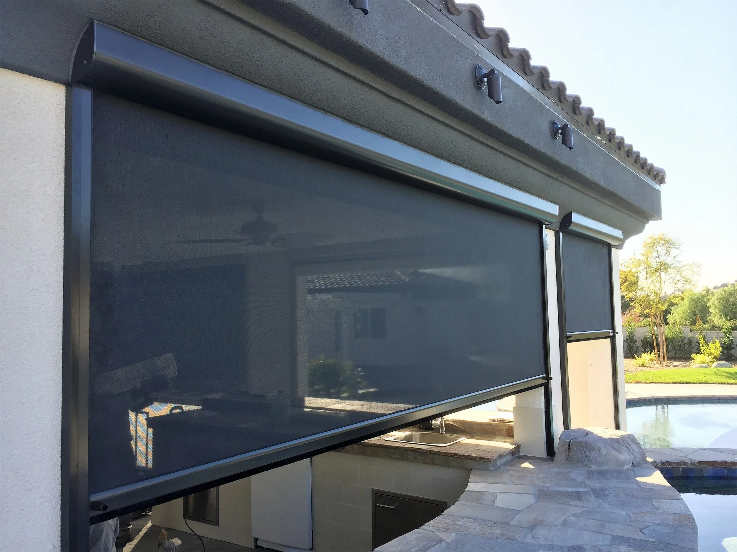 Outdoor view of a modern building with a large black retractable screen and a small outdoor sink, surrounded by trees and a clear sky.