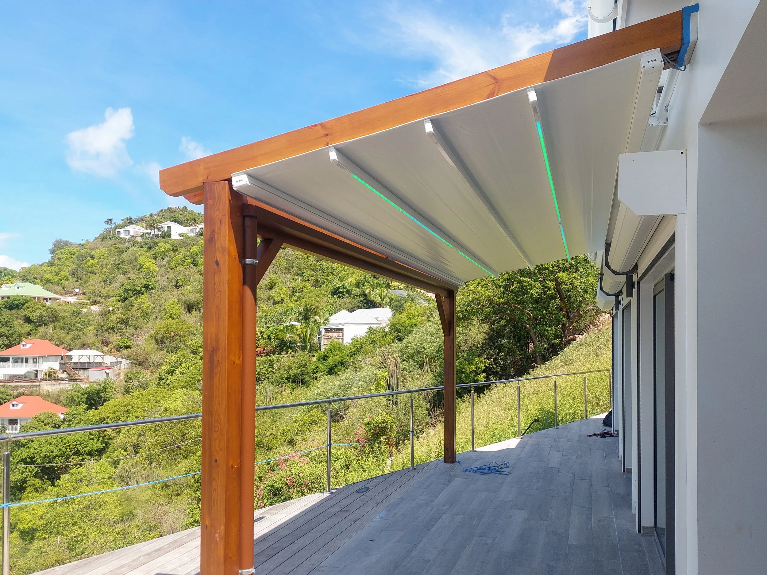 A patio with a wooden pergola and white retractable awnings, overlooking a lush green hillside with houses.