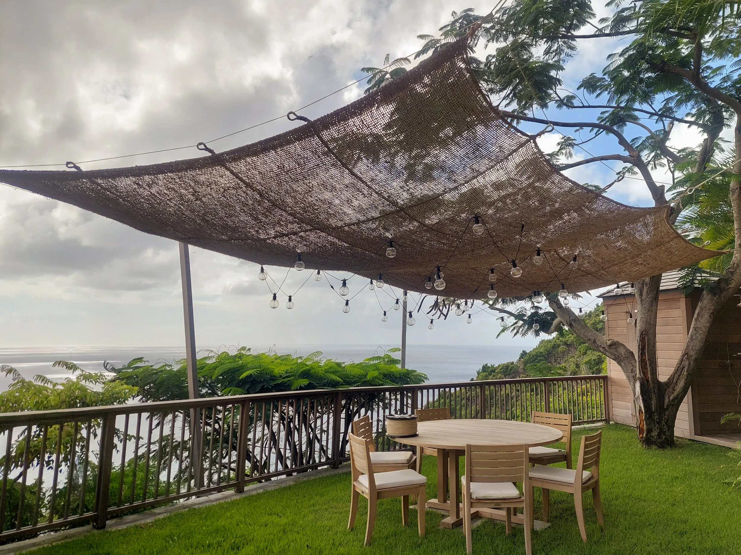 Outdoor patio with wooden table and chairs, surrounded by greenery and overlooking the ocean, with a woven canopy overhead and string lights.