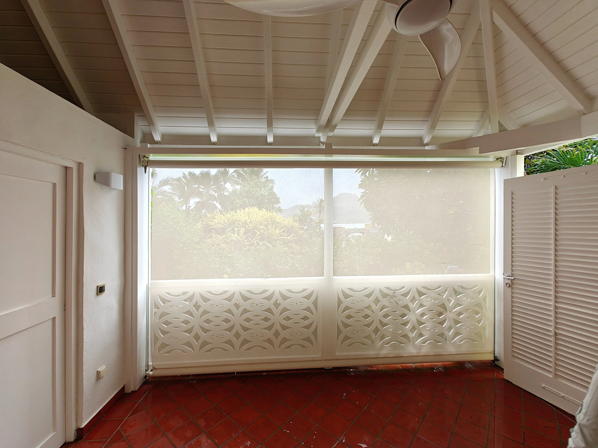View of a screened porch with a white ceiling, a decorative white lattice privacy wall, and red tiled flooring. The porch faces lush green trees and blue sky outside.