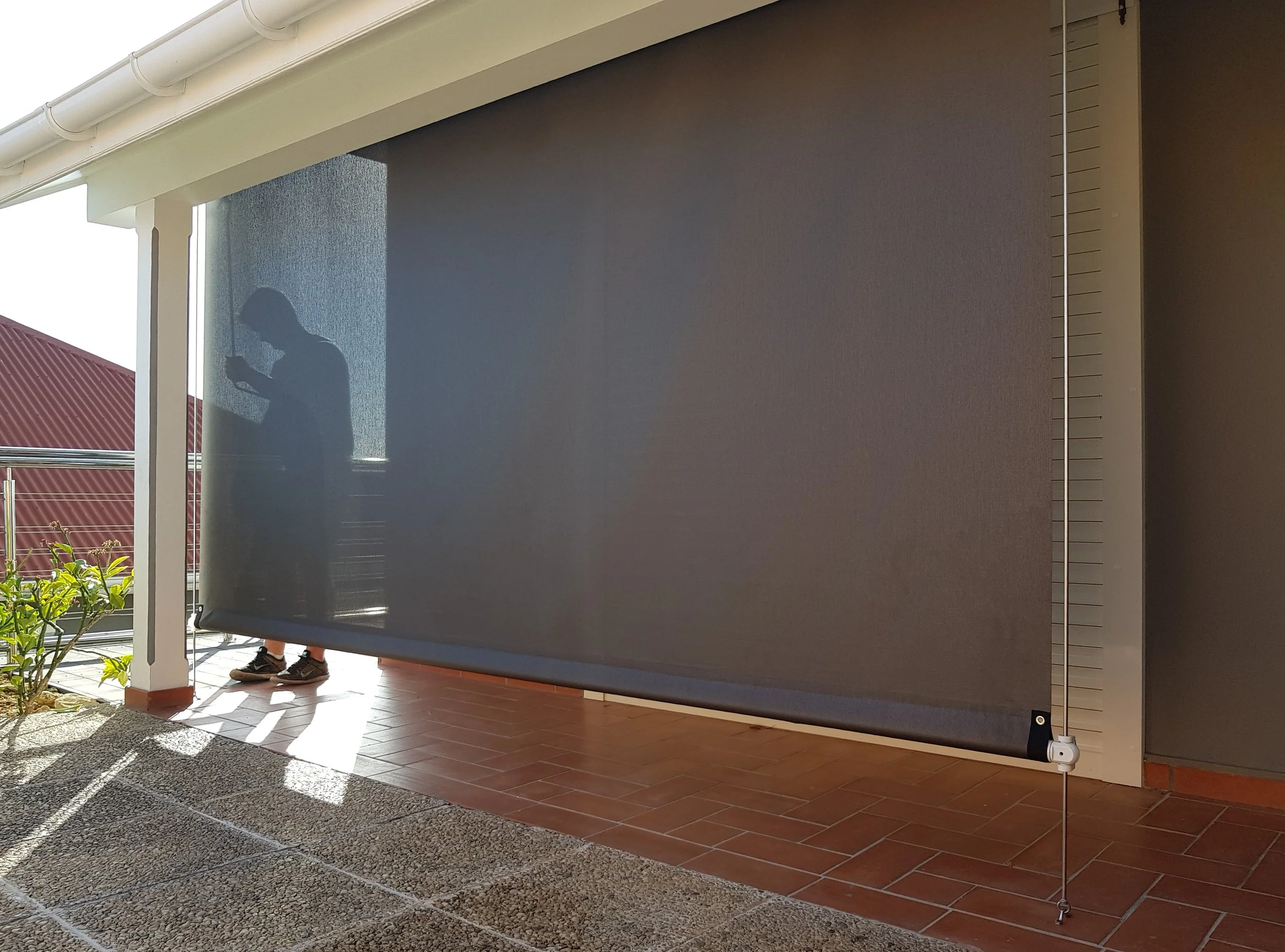 A person with dark pants and sneakers standing next to a large gray outdoor roller shade or screen, which is partially rolled down, on a porch or patio with tiled flooring. There is a white gutter pipe running along the roof edge and some greenery in