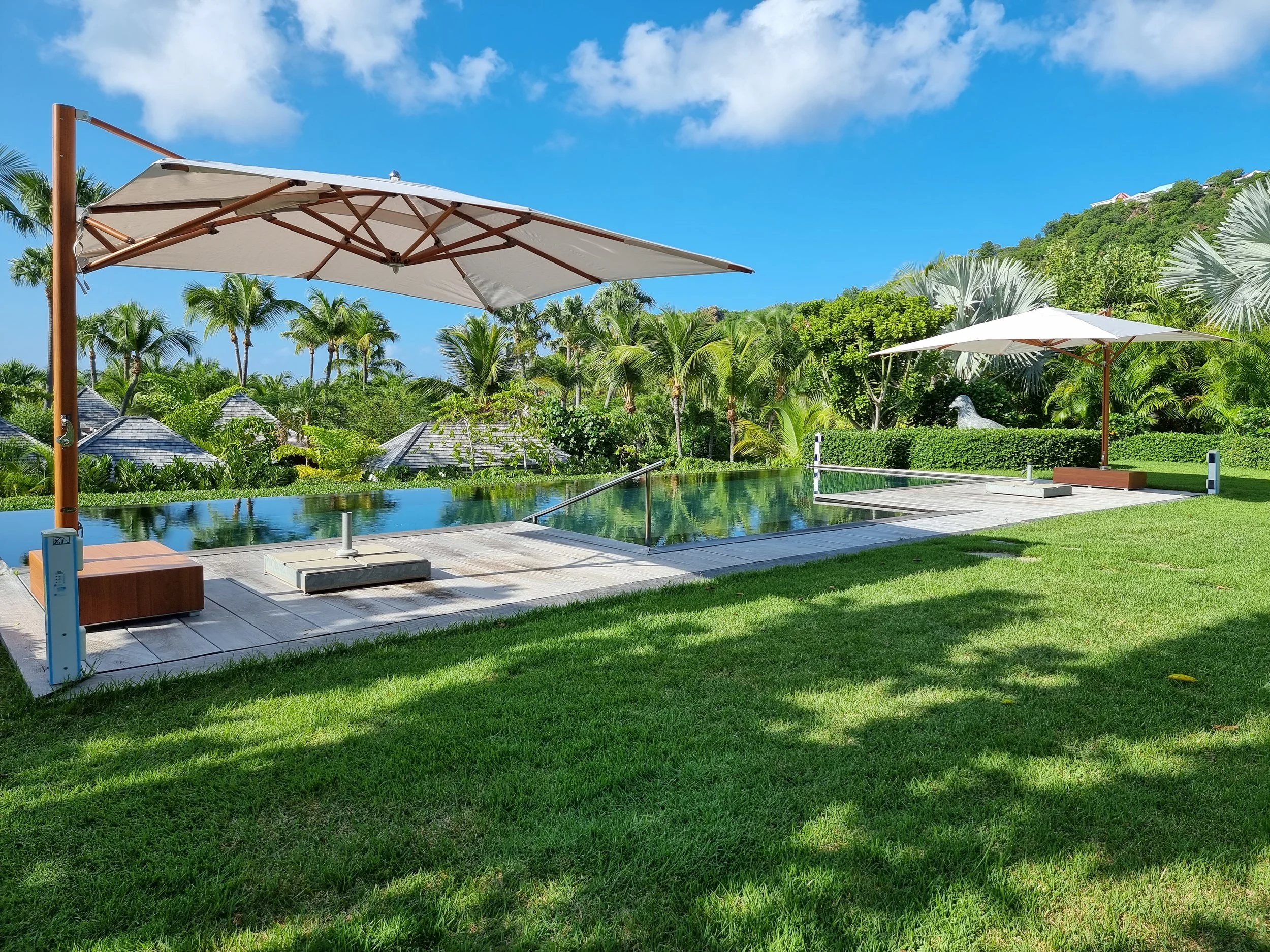 A tropical outdoor setting featuring a swimming pool with two white patio umbrellas, lush green trees, and a bright blue sky with scattered clouds.