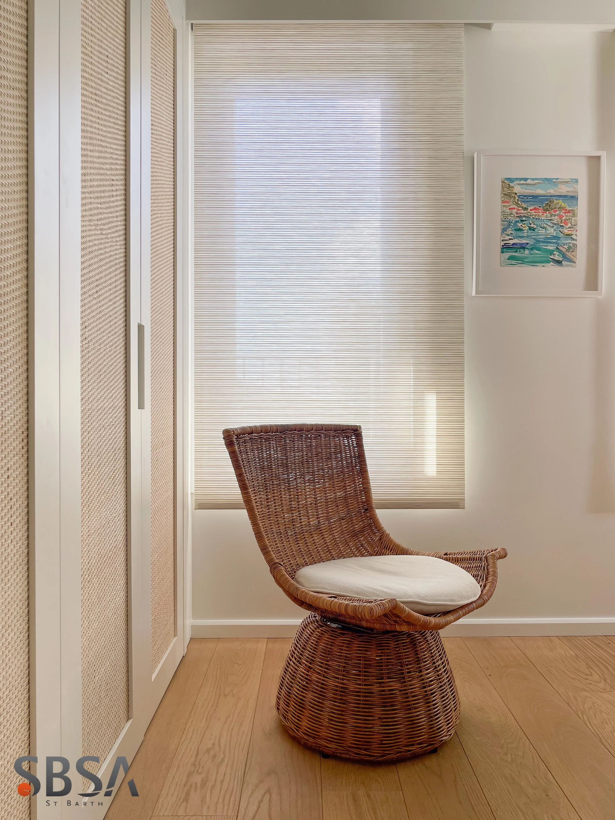 A wicker chair with a cushion in a room with wooden flooring, a window with horizontal blinds, a picture frame on the wall, and a cabinet with woven doors.