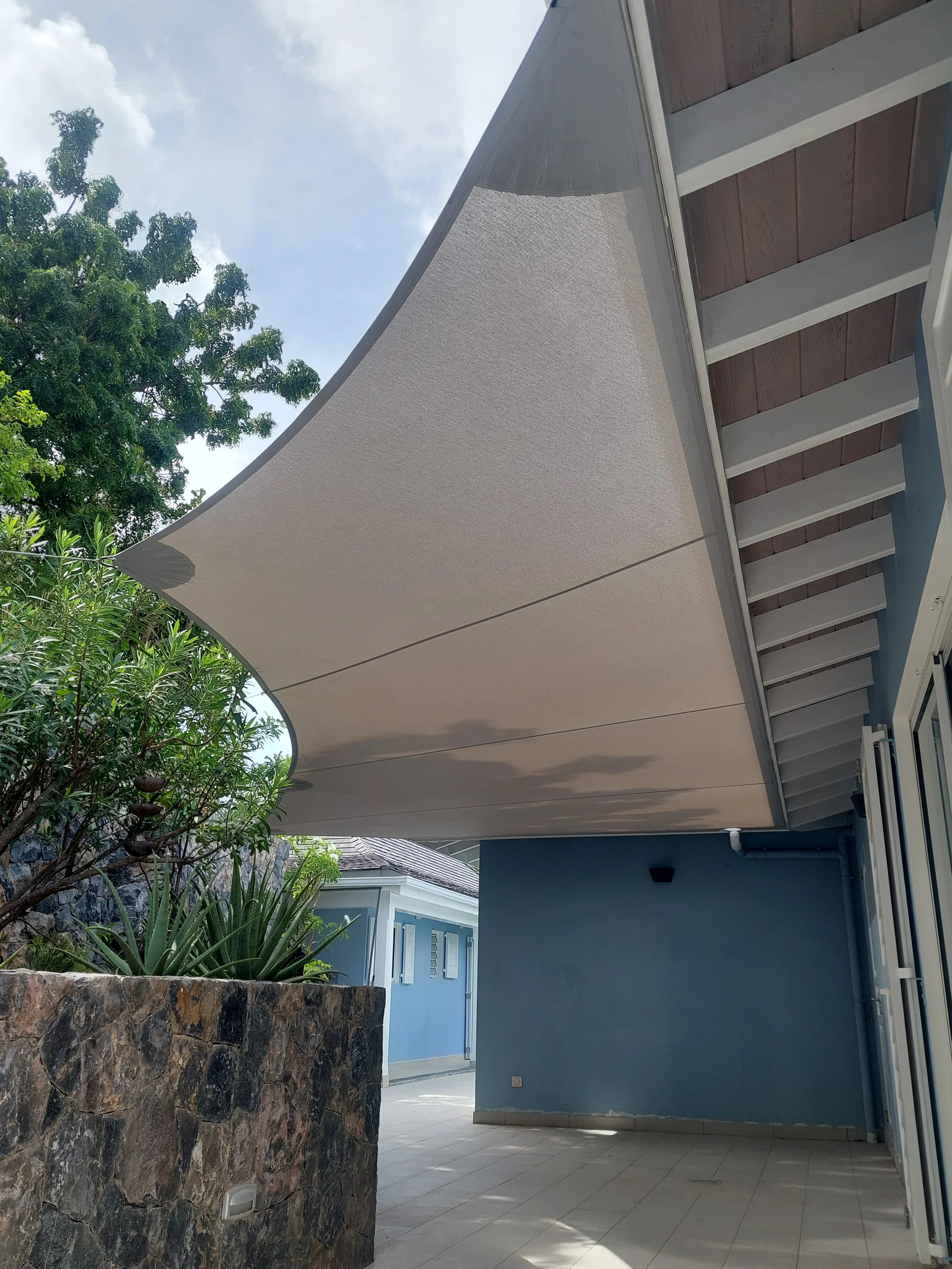 An outdoor patio area with a beige awning, a stone wall with plants, a blue building with a window, and a tiled floor.