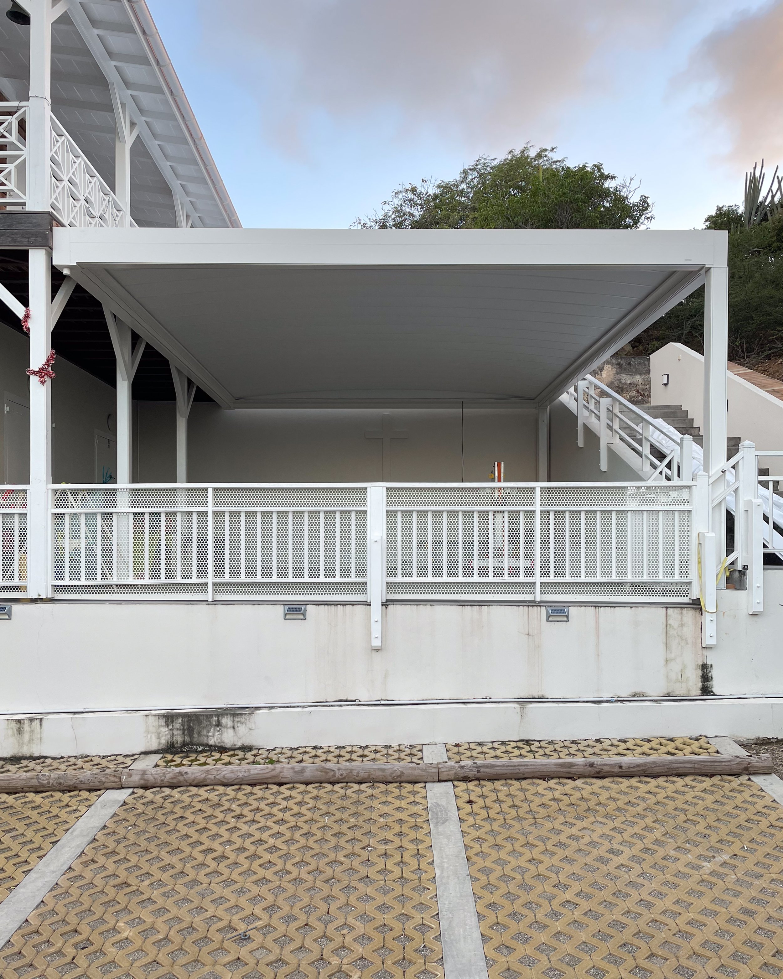White retractable pergola elevated on a large terrace. Large metallic white structure and a parking lot in front.