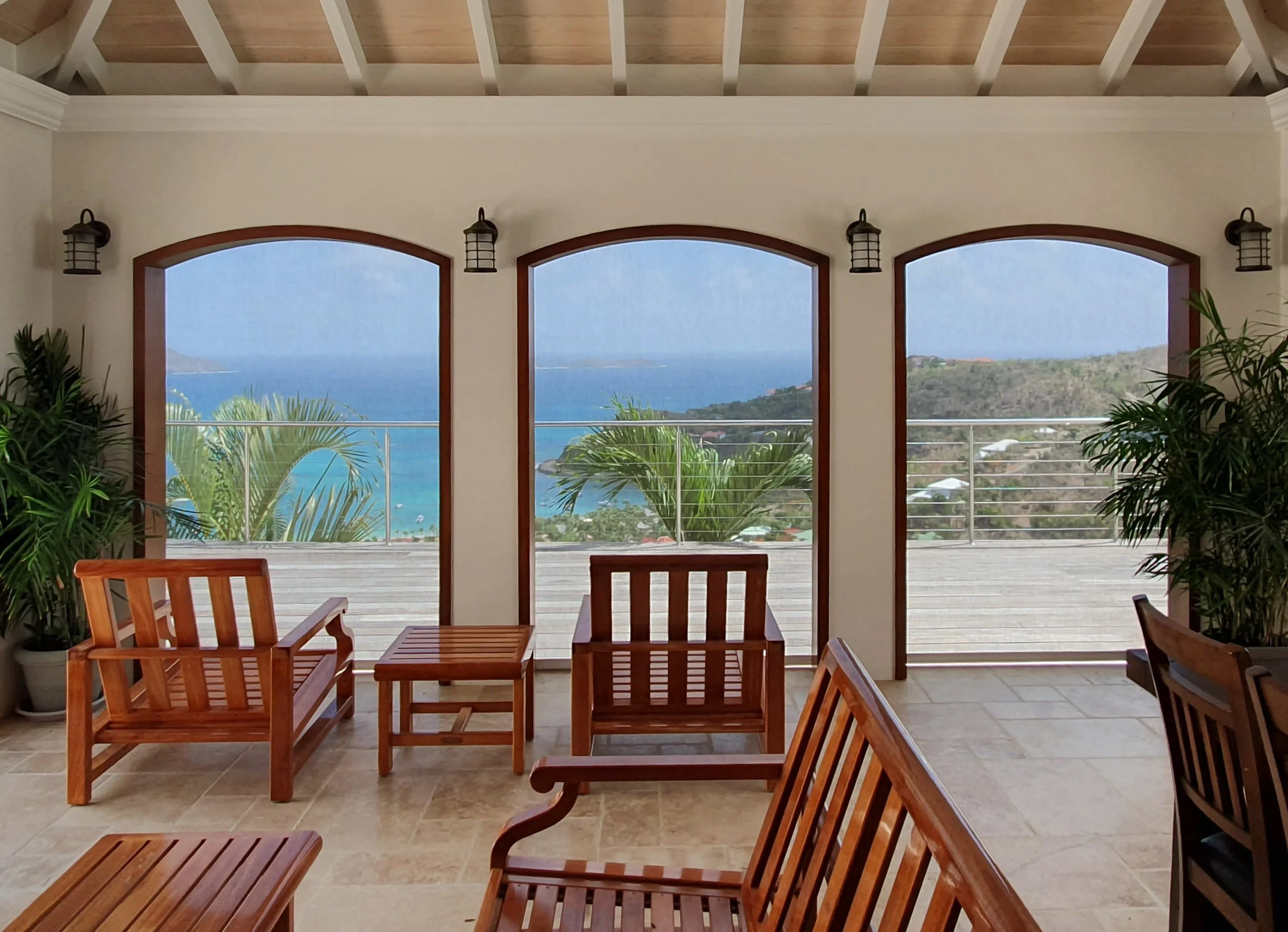 View of the ocean with hills and houses in the background, seen through large arched windows in a room with wooden chairs and indoor plants.