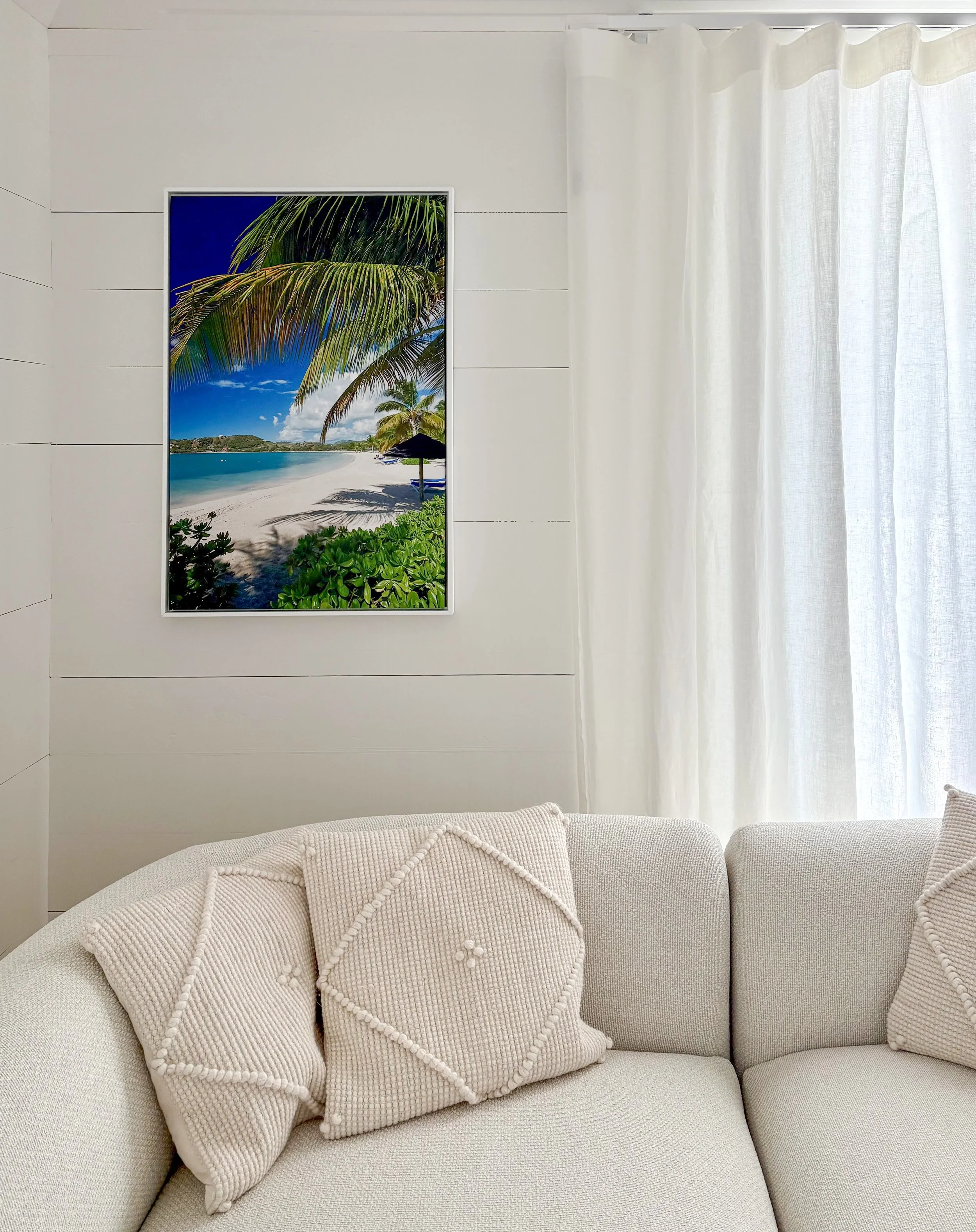 Inside a light-colored living room, a white sofa with textured, geometric pillows is visible. On the plain, white paneled wall behind, there is a framed large photograph of a tropical beach scene with palm trees, sand, and calm ocean water. To the right, sheer white curtains cover a window, allowing natural light to fill the room.