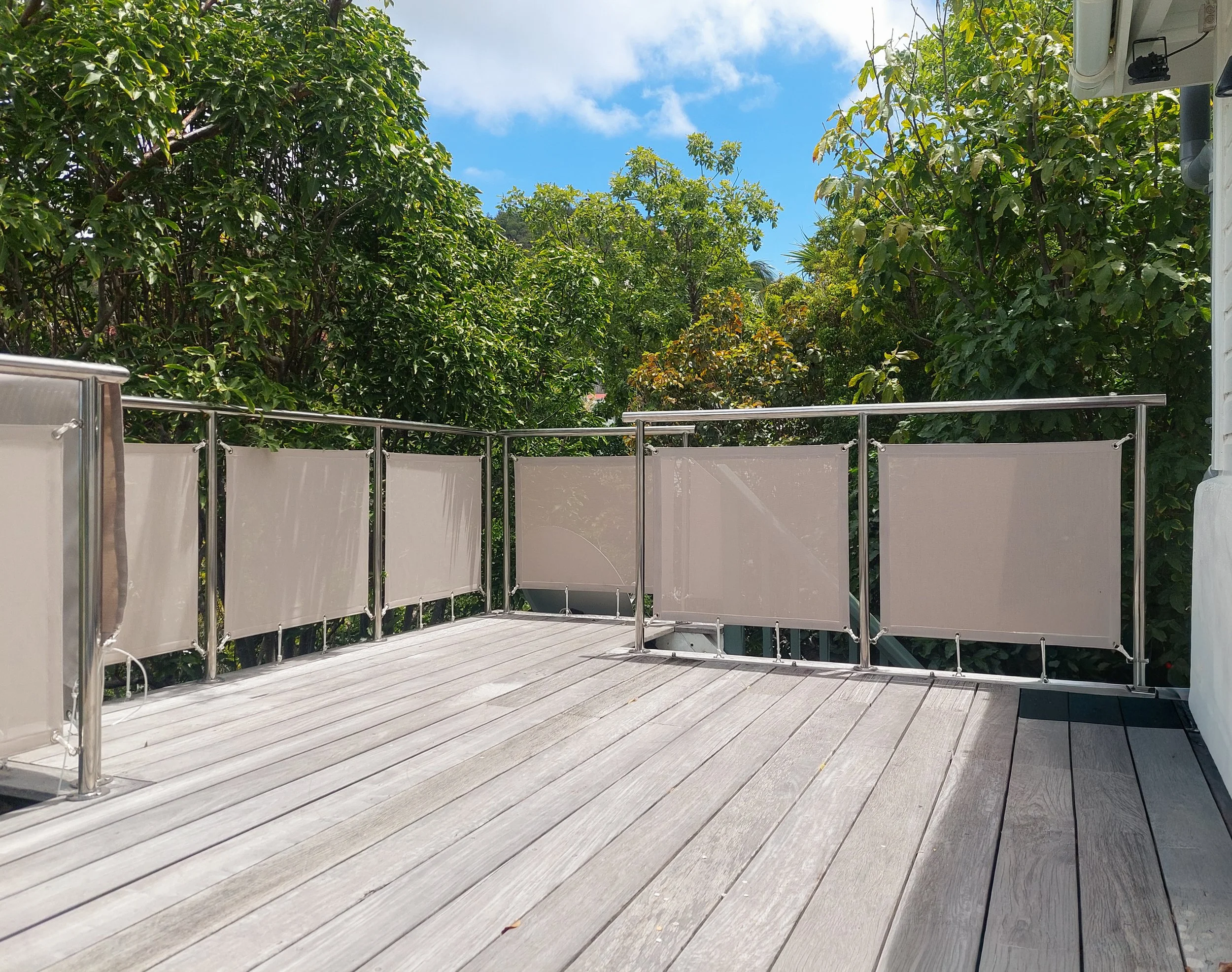 An outdoor wooden deck enclosed by a rope and fabric barrier, surrounded by green trees and bushes under a blue sky with white clouds.
