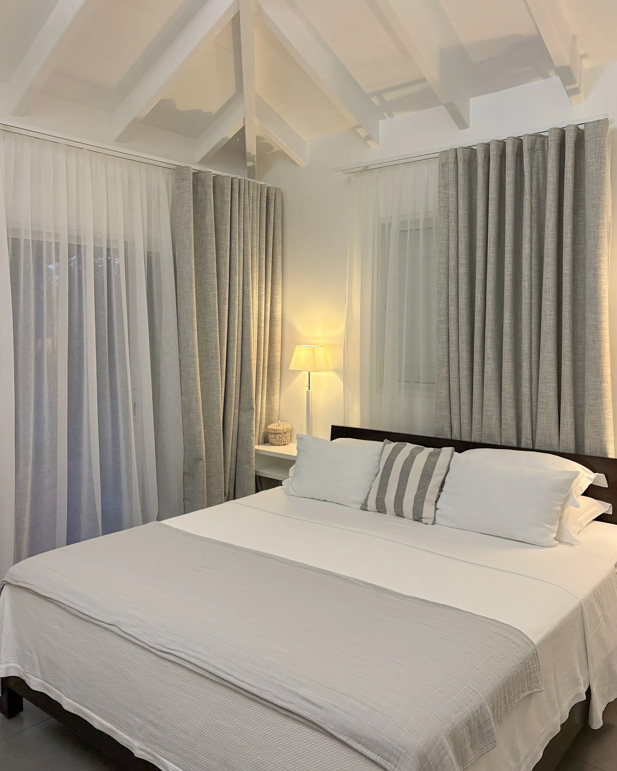 A neatly made bed with white bedding and striped pillows in a bedroom with beige curtains and a bedside lamp.
