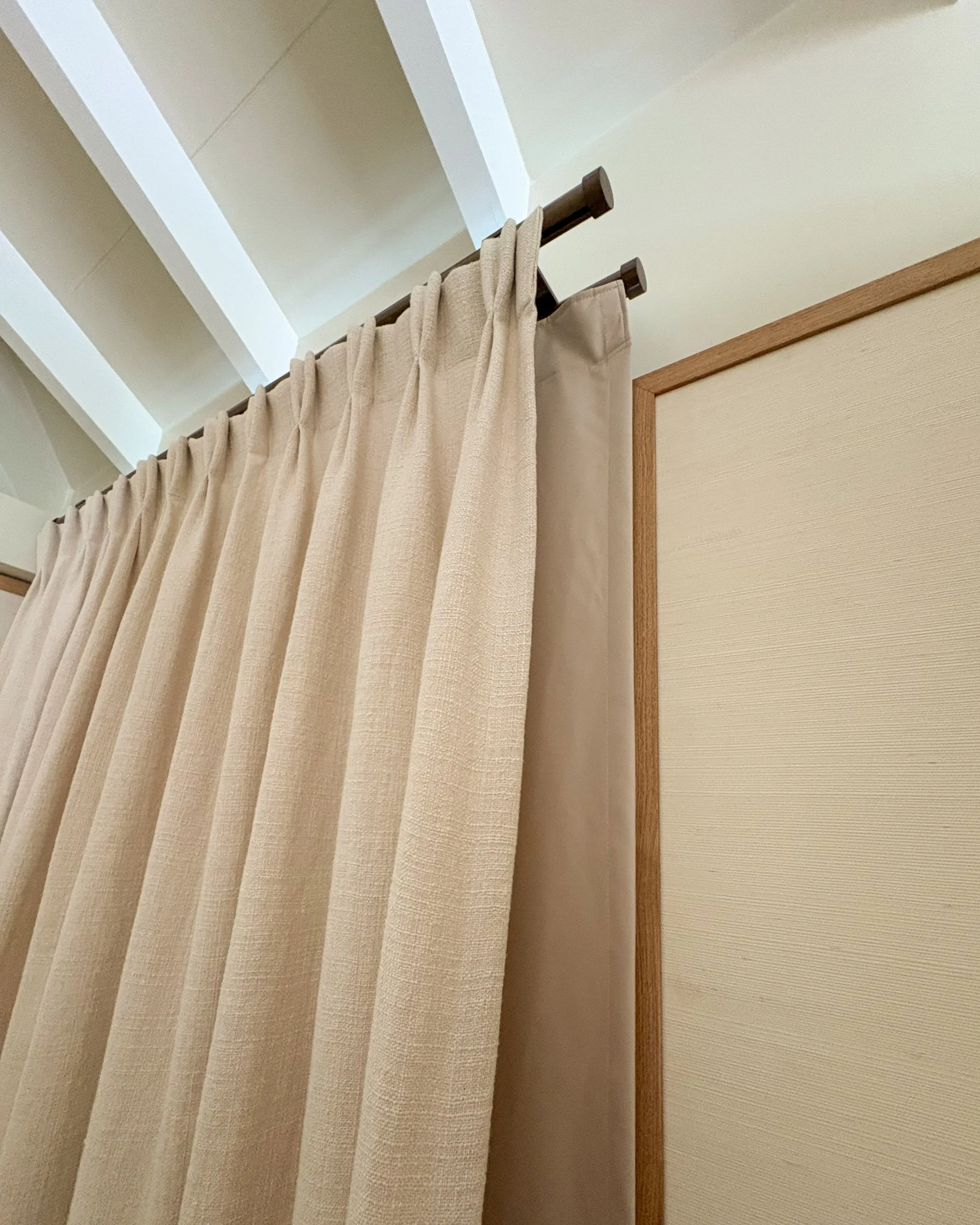 A beige curtain hanging from a dark brown curtain rod in front of a beige textured wall and ceiling with exposed beams.