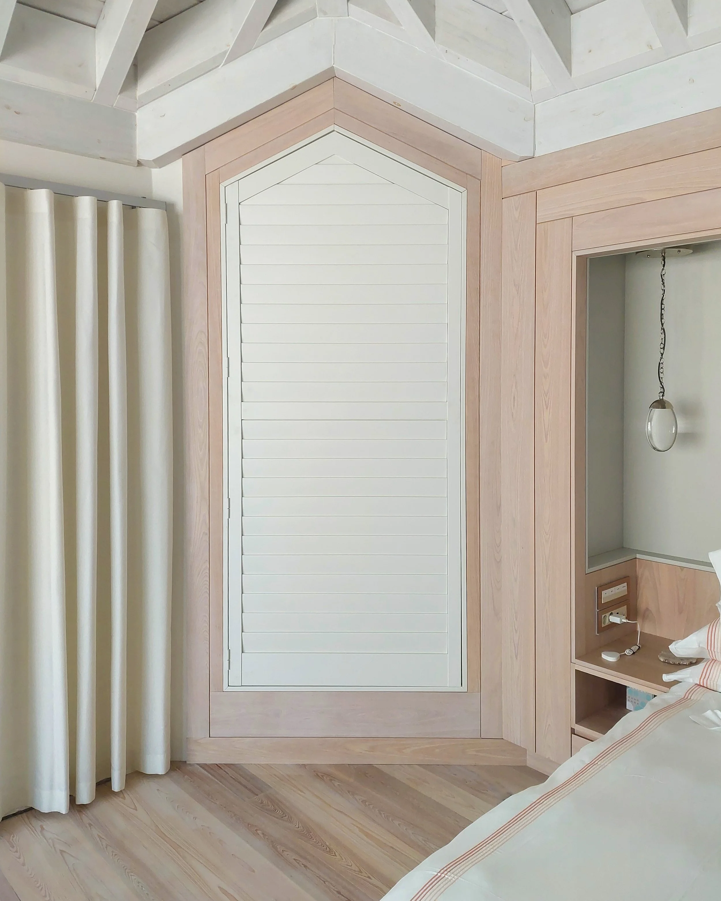 Un coin de chambre montrant une partie d’un lit avec un oreiller à rayures, une table de chevet en bois avec une prise électrique, un mur blanc avec une fenêtre habillée de rideaux blancs, ainsi qu’un mur d’accent en bois doté d’une fenêtre à persien