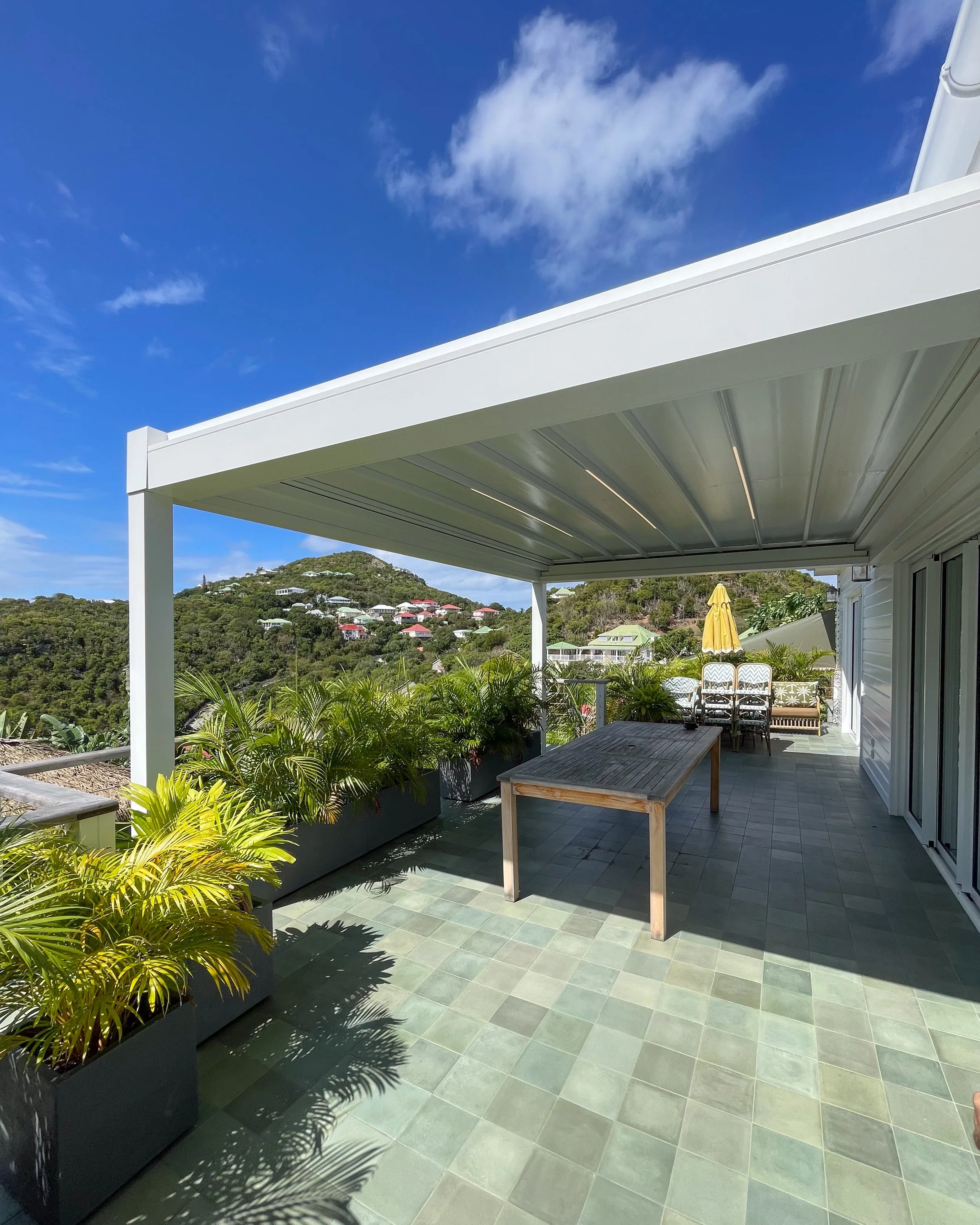 A patio with a white pergola, potted tropical plants, a wooden table, and stacked chairs. In the background, there are hills with houses, a clear blue sky with clouds.