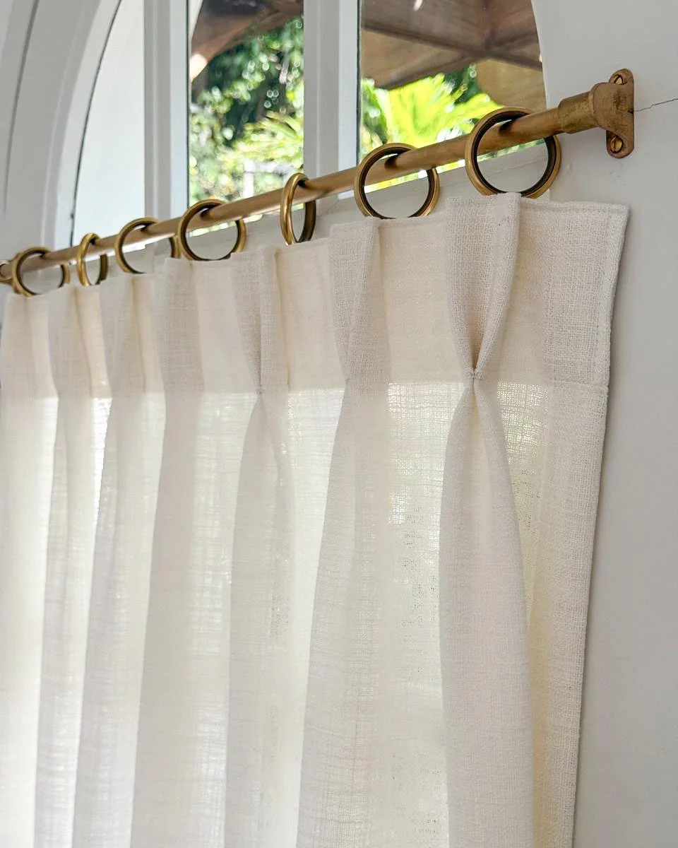 Cream-colored sheer curtains hanging on a brass curtain rod with rings, in front of a window with greenery outside.