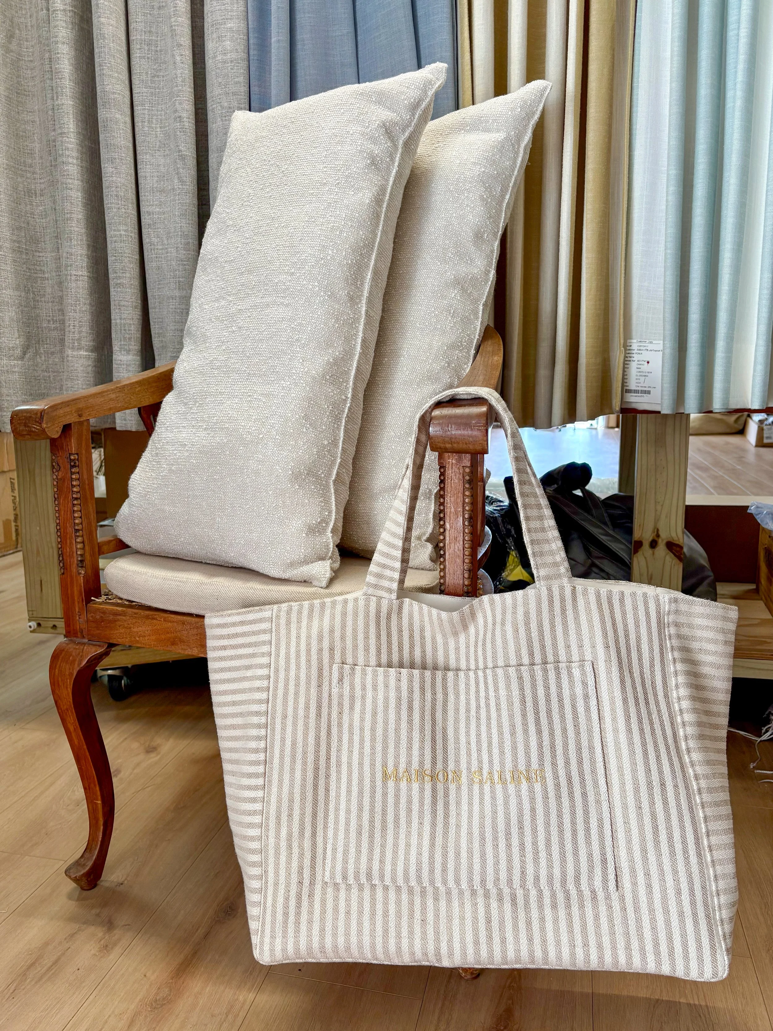 A display of fabric and curtain samples on a wooden fixture in a store, with a tote bag labeled 'Maison Salint' hanging from the chair.