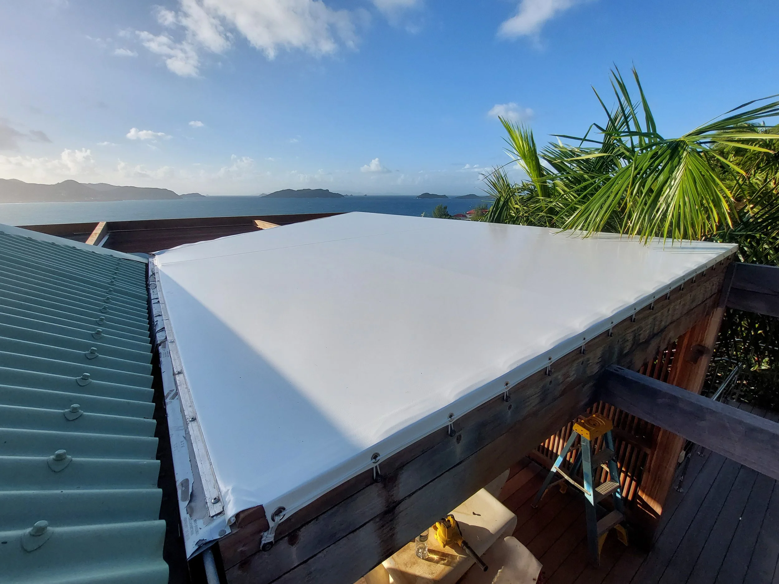 Roof with green and white panels, overlooking a coastal landscape with hills, ocean, and palm trees, on a clear sunny day.