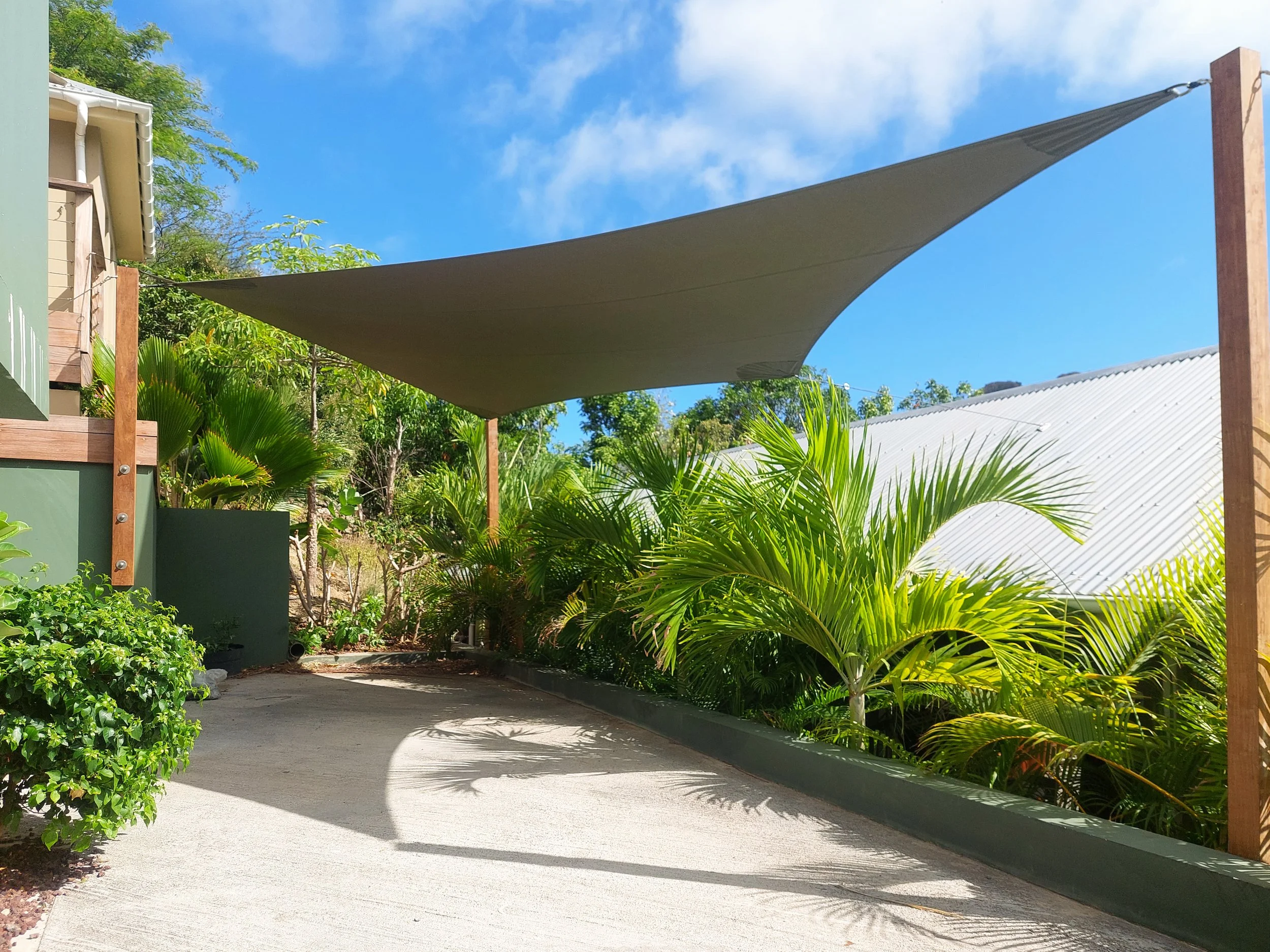 A beige fabric shade sail is suspended above a concrete pathway in a garden area, supported by wooden posts. The garden is lush with green plants including small trees and shrubs. The sky is blue with some scattered clouds.