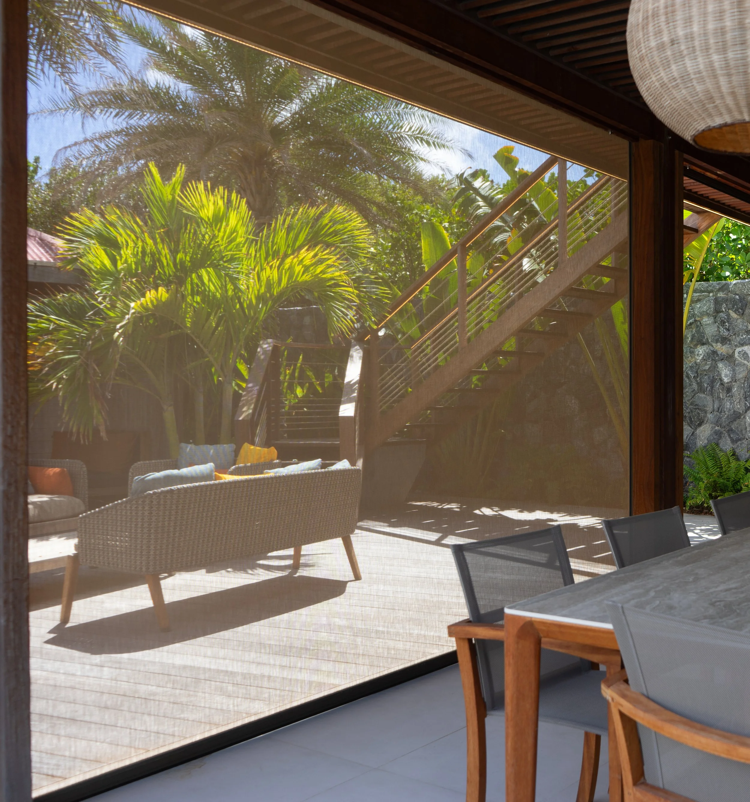 View through a glass door showing a wooden deck with outdoor furniture, surrounded by lush green tropical plants and trees, with sunlight illuminating the scene.