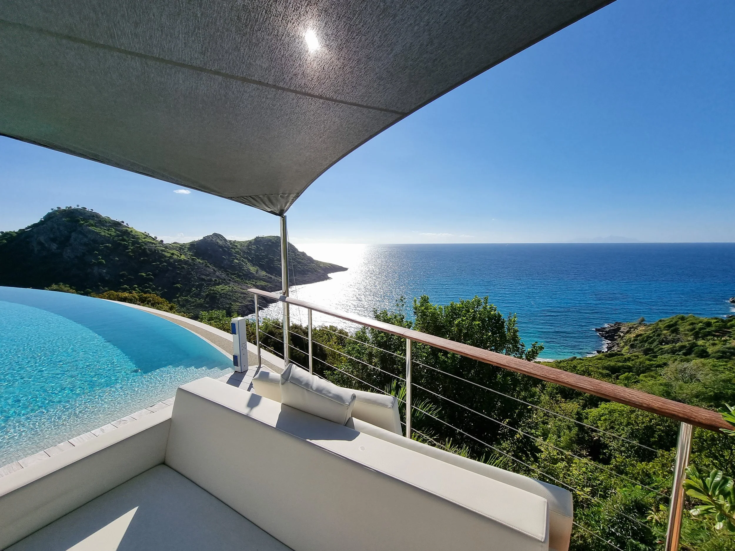 A view of the ocean and green hills from a balcony with a curved lounge chair, overlooking a swimming pool, under a large shade umbrella.