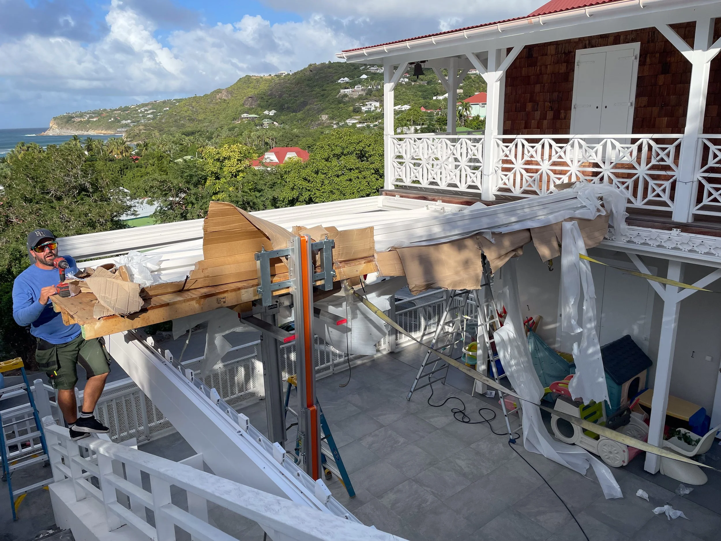 Construction workers installing a retractable pergola beside a house with a view of a hillside and ocean in the background.