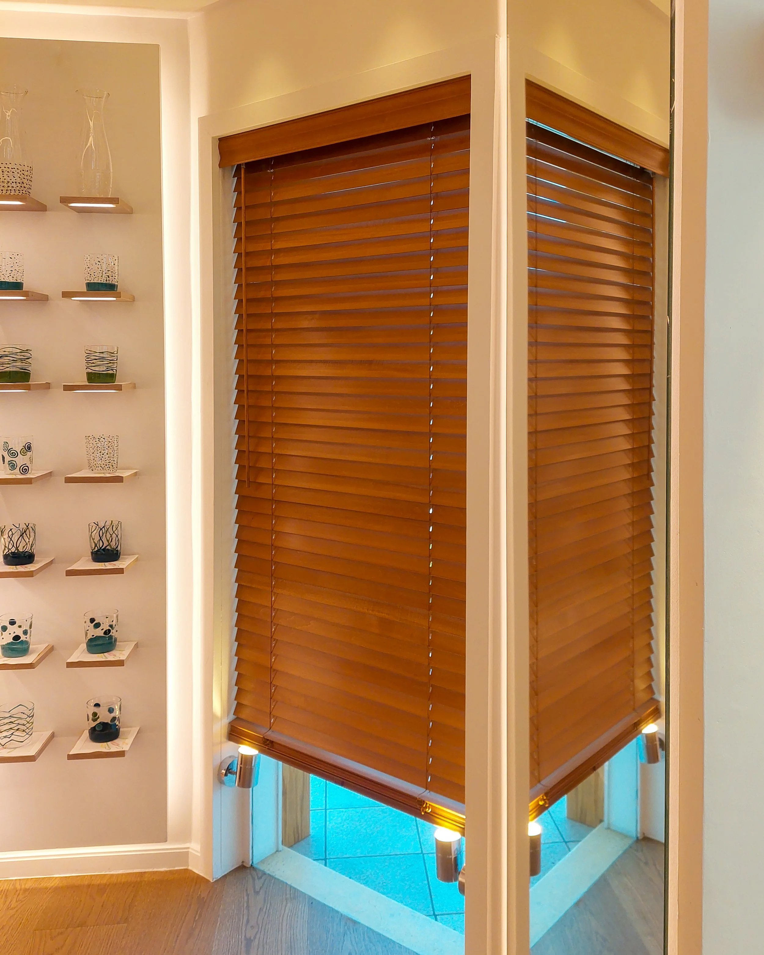 Vue intérieure d’une pièce avec des stores horizontaux en bois sur une fenêtre, un escalier bordé de bougies sur le côté, ainsi qu’une étagère décorée de vases en verre et de petits contenants en céramique.